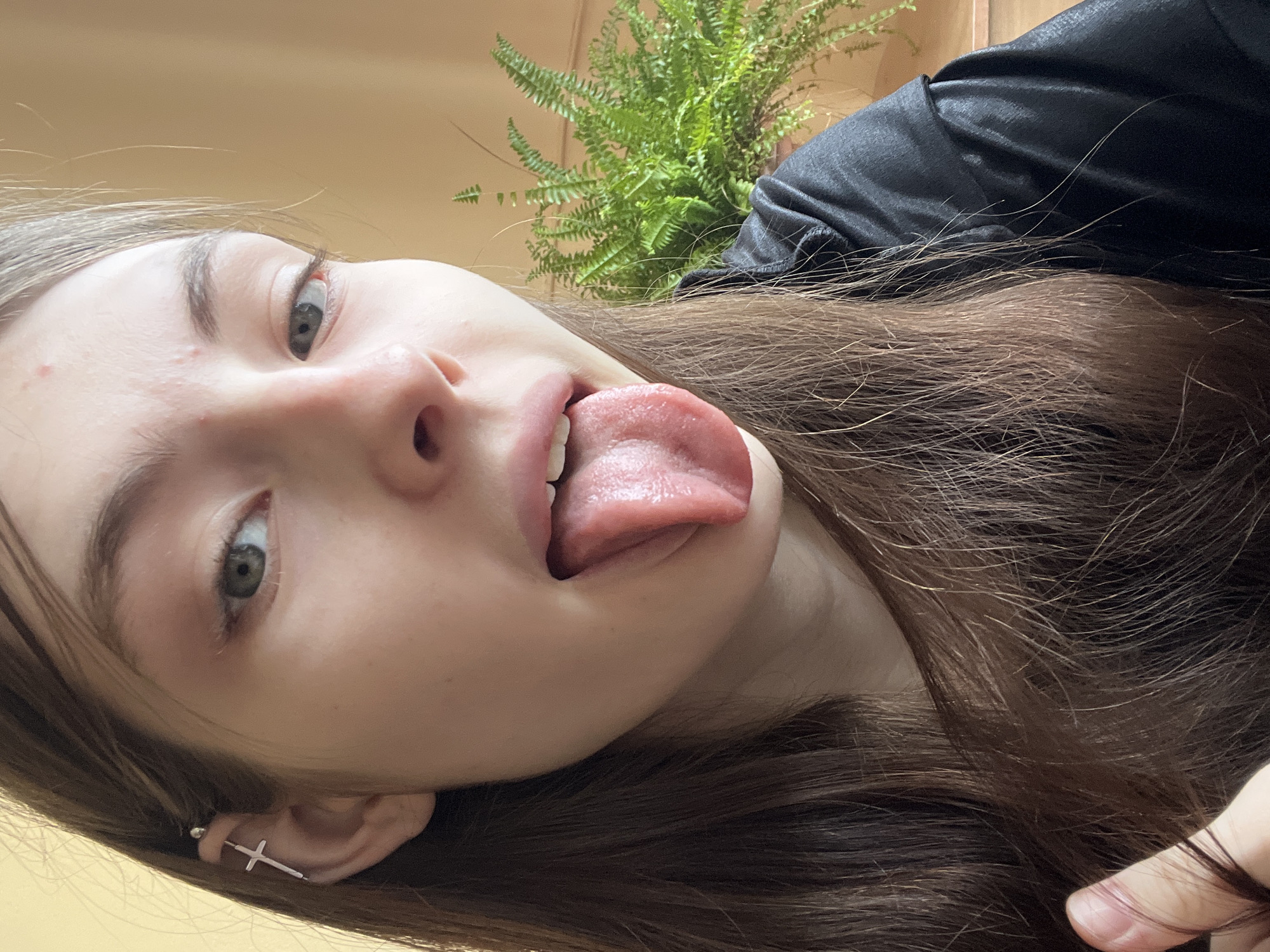 A close-up, tilted portrait features a young person, likely female, captured indoors in Saint Petersburg, Russia. The subject has long, straight brown hair that frames their face and extends into the bottom right corner of the frame. Their fair skin shows some minor blemishes on the forehead. The person has expressive green-grey eyes, looking directly at the camera with a playful expression. Their tongue is sticking out of their mouth.
On the left side, a silver cross-shaped earring is visible in the earlobe, along with another silver barbell-style piercing higher on the ear. The background consists of a plain, light brown or beige wall. In the upper right portion of the image, a vibrant green fern plant is partially visible, indicating an indoor setting. The subject is wearing a black garment, possibly a shirt or jacket, with visible fabric folds. A hand with neatly manicured fingernails is seen gently touching a section of hair in the bottom right. The lighting suggests it is daytime or a well-lit indoor space. No text is visible in the image.