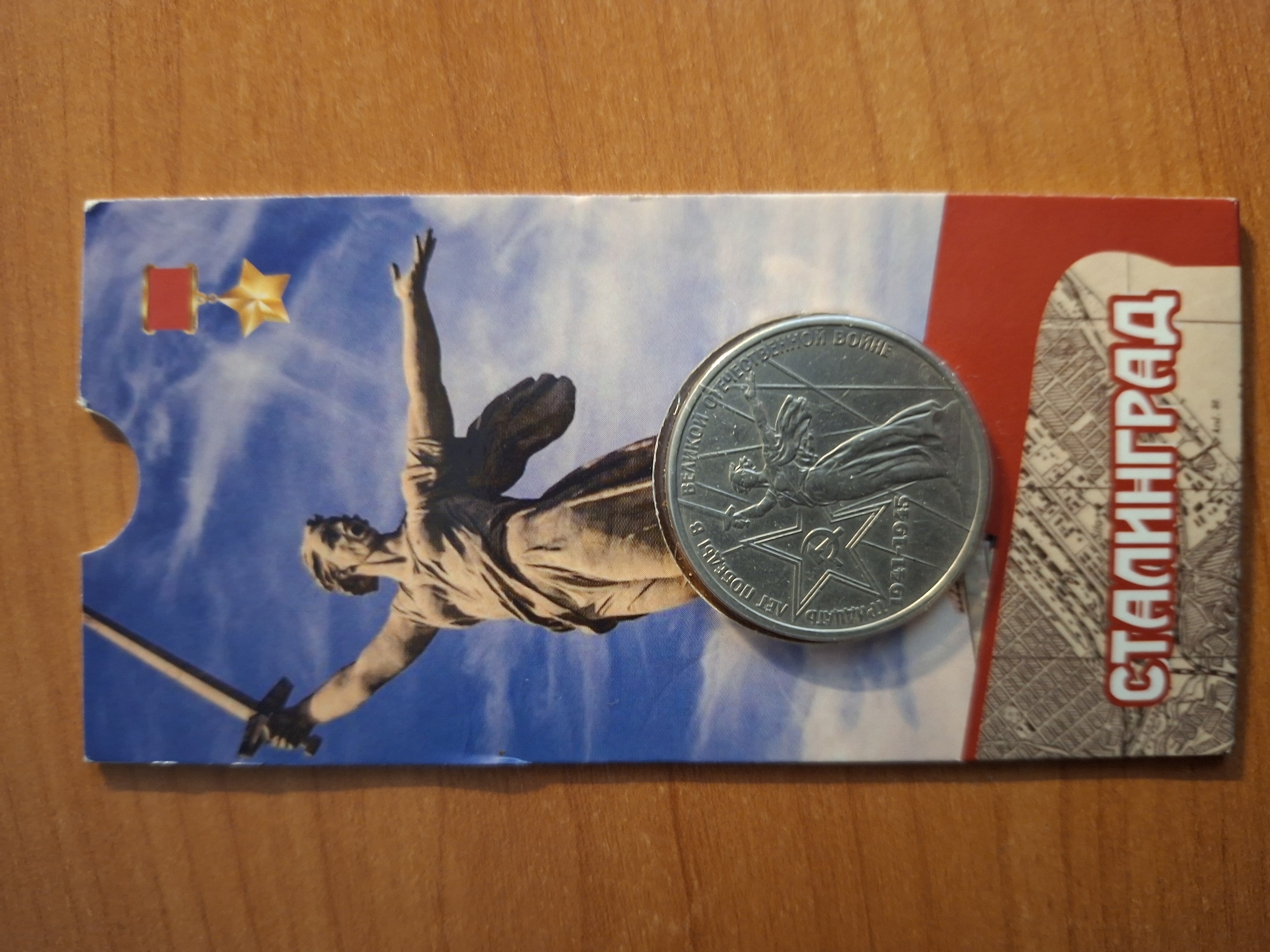 The image shows a postcard with a depiction of the Motherland Calls statue in Volgograd, Russia, and a commemorative coin. The statue, a monumental work of art and a symbol of Soviet victory in World War II, stands tall against a dramatic blue sky with scattered clouds. The coin, placed in the foreground, is silver and features a relief of the statue on one side, surrounded by inscriptions and the years 1941-1945, indicating its connection to the Great Patriotic War. The postcard also features the word "СТАЛИНГРАД" (Stalingrad) in large, bold letters at the bottom, a historical name for the city, emphasizing its significance in the war. The postcard is lying on a wooden surface, likely a table, and appears to be a souvenir item.
