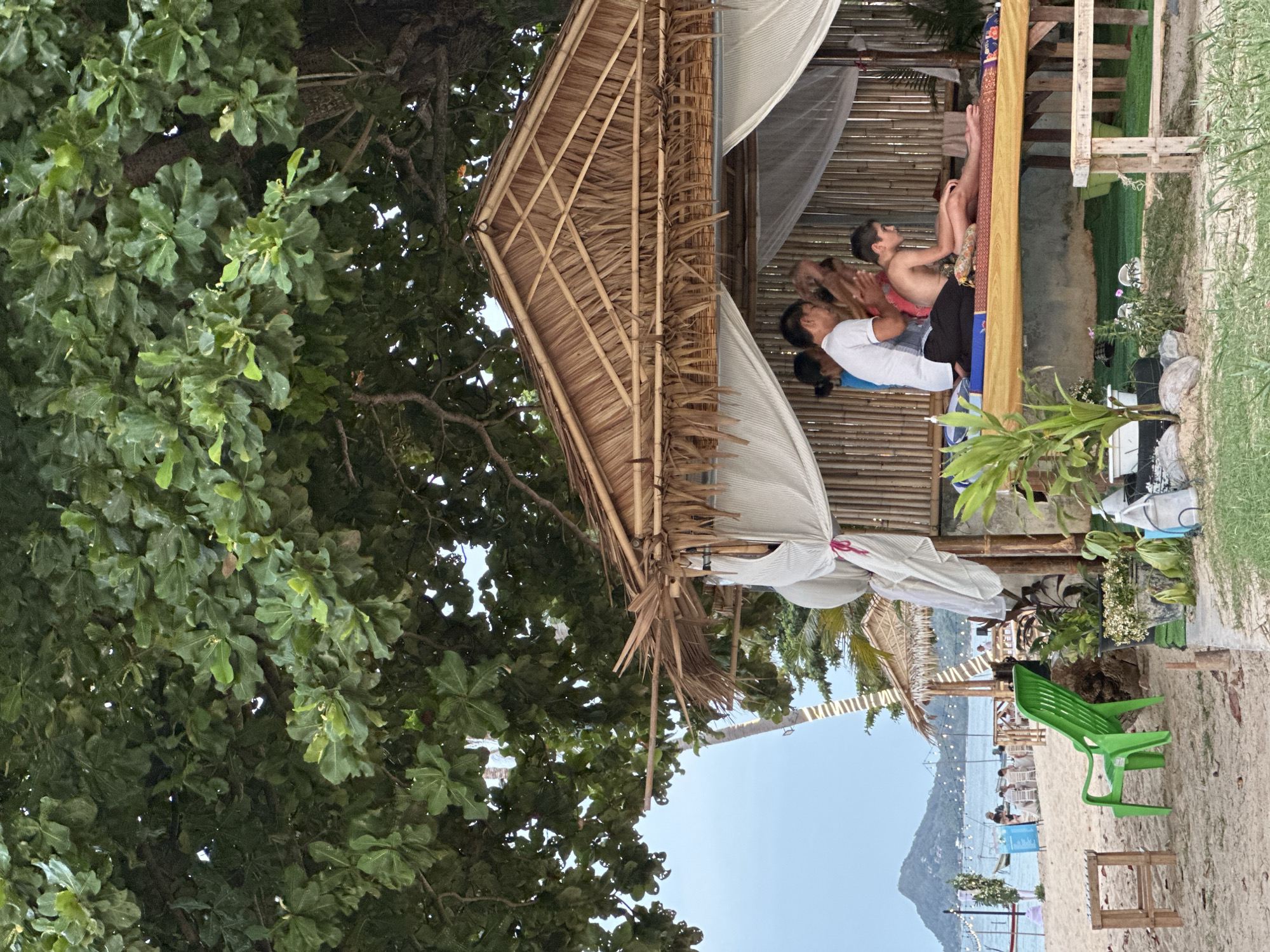 This image depicts an outdoor massage service on a beach in the Ko Samui District of Thailand. Underneath a large, leafy tree, a thatched-roof hut with bamboo walls provides shelter for a massage setup. Two massage therapists are attending to a young male client lying on a mat on a raised wooden platform. White sheer curtains hang around the open sides of the hut. In the background, the beach is visible, with people seated at tables further down the shore and a calm ocean with a distant island. A bright green plastic chair is positioned on the sand in the foreground, along with a small wooden stool and potted plants. The overall scene conveys a relaxed atmosphere typical of a tropical beach resort.