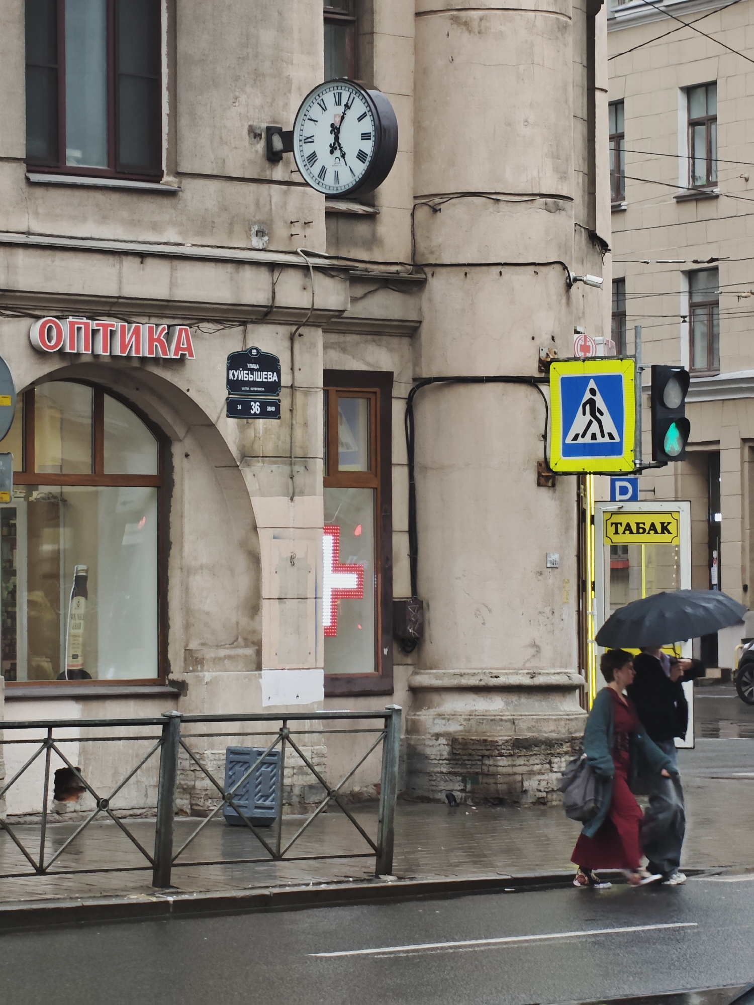 The image depicts an exterior view of a building in a city, with a street and sidewalk in the foreground. A clock is mounted on the side of the building, showing the time as approximately 5:10. A red neon sign with the Cyrillic word "ОПТИКА" (Optics) is visible above a shop window. A street sign displays "УЛИЦА КУЙБЫШЕВА 36" (Kuibysheva Street 36). A pedestrian crossing sign is present with a blue background and white pedestrian symbol. A traffic light shows a green signal. Two people are walking across the street under a black umbrella. The ground is wet, indicating that it is raining. A metal fence runs along the sidewalk.