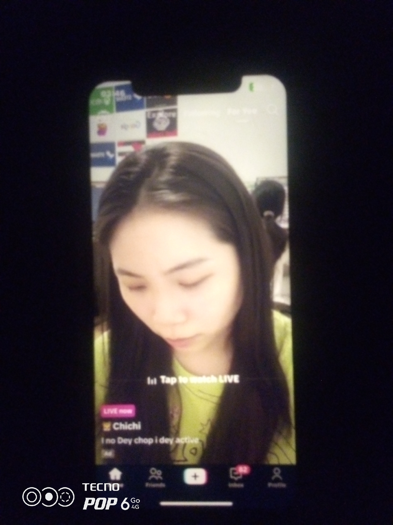A medium shot captures a young woman with dark hair, looking down with her eyes partially closed, as if she is concentrating or about to speak. She is wearing a bright yellow shirt. The image appears to be a screenshot from a mobile phone, as indicated by the interface elements visible at the bottom and top of the screen. A "LIVE now" banner is displayed, along with the text "Chichi" and "I no Dey chop i dey active." Other icons and text are also visible, suggesting a social media or live-streaming application. The background behind the woman is blurred, but it seems to depict a wall with various logos and images. The overall lighting suggests an indoor setting. The lower portion of the screen displays "TECNO POP 6 4G" branding. The image is not from Howayi, Nigeria.