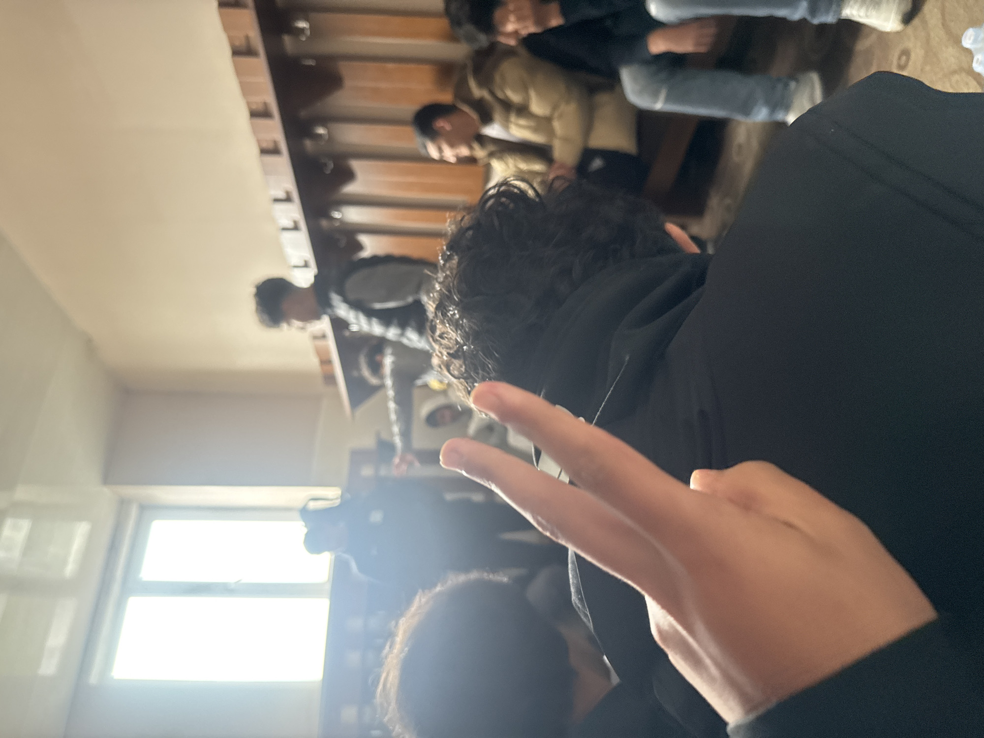 A cluttered indoor scene, likely a changing room or lounge area, is depicted with a tilted perspective. Multiple people are present, including several seated on wooden benches, identifiable by visible backs, heads, and partial torsos. These individuals appear to be wearing a mix of casual clothing, including jackets. One person is in the foreground. The individual is also wearing a black hoodie and is making a V-sign with their fingers. Windows in the background provide a source of strong backlight. Based on the instructions, this picture was probably taken in the city of Charentsavan, Armenia.