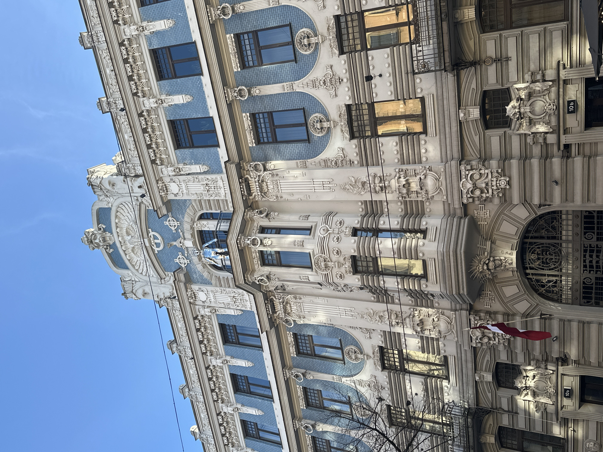 The image shows a close-up of the facade of an Art Nouveau building in Riga, Latvia. The building has a blue and white color scheme, with ornate decorations including relief sculptures, pilasters, and window frames. Windows are set in a grid pattern with brown frames. Sunlight reflects in some of the windows. A Latvian flag is attached to the building. A sign reading "10b" is visible. A tree with bare branches is in the foreground. The sky is blue.
