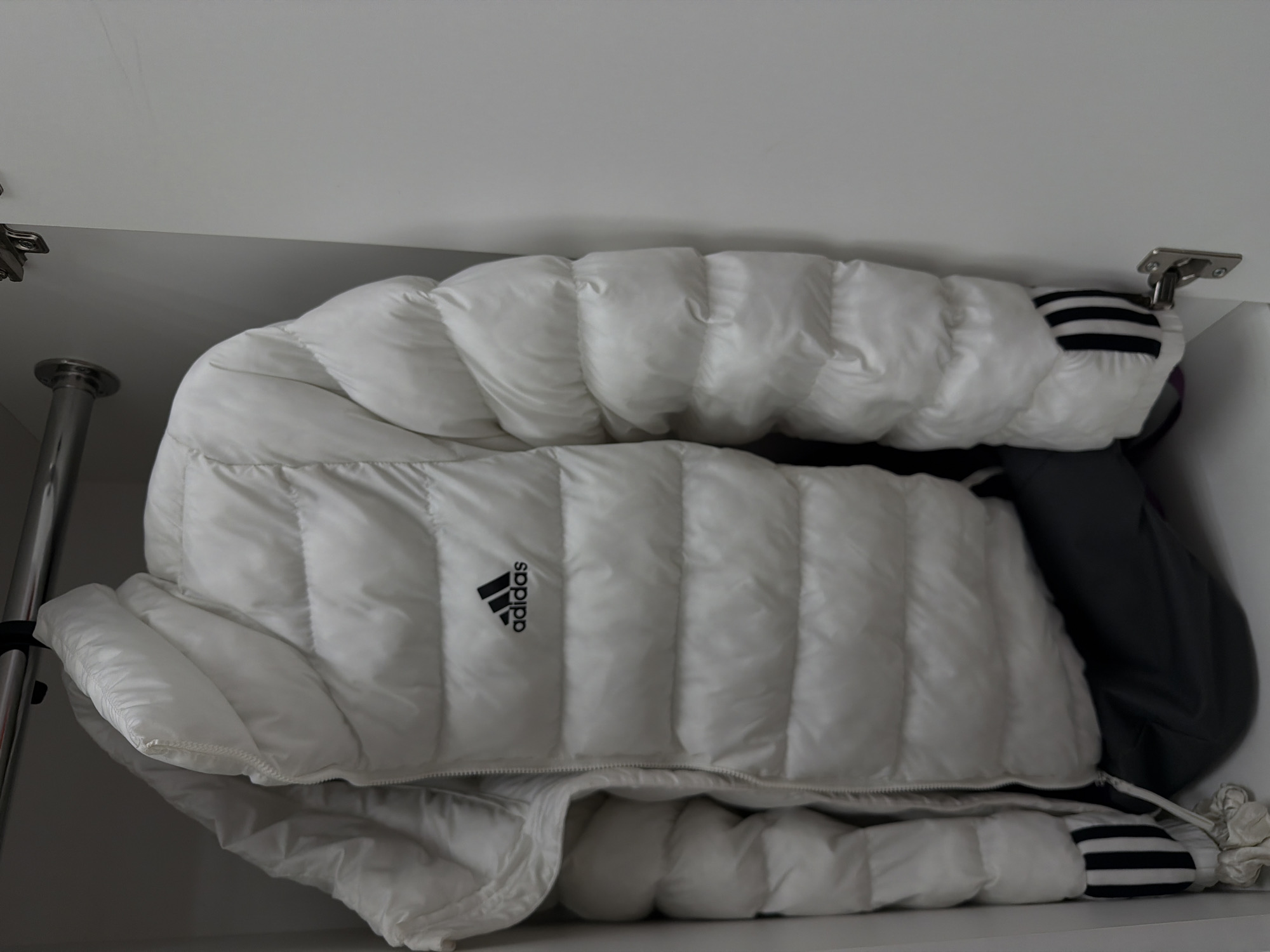 A folded white Adidas down jacket rests in a white closet, likely in a residential setting, though its specific location in Izhevsk, Russia, is indeterminable from the image. The jacket is quilted and features the Adidas logo on one side. A gray garment with striped sleeves is partially visible alongside the jacket. The closet contains a metal clothes rod and hinges. The overall lighting is soft and diffused.