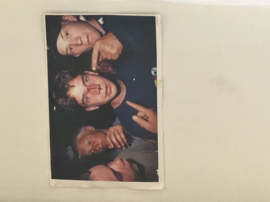 The image displays a physical color photograph, partially affixed to a light-colored surface, depicting four young adult males in a dimly lit setting.
In the center, a male with short brown hair, wearing a dark blue t-shirt, has a prominent bloodstain on his nose and upper lip. His left hand is raised in a gesture with the index and pinky fingers extended. He gazes directly at the viewer.
To his immediate right, another male, wearing a dark cap, extends his right index finger, pointing directly at the central male's bloody nose.
To the central male's left, a male with light-colored hair holds a clear glass or bottle containing liquid. Partially visible on the far left is a fourth male, wearing a grey t-shirt, observing the scene.
The background is dark and indistinct, providing no discernible environmental details. No visual information within the photograph allows for the determination of a specific location in Paris, France, or any other geographical area. The overall scene appears to be a casual group snapshot.