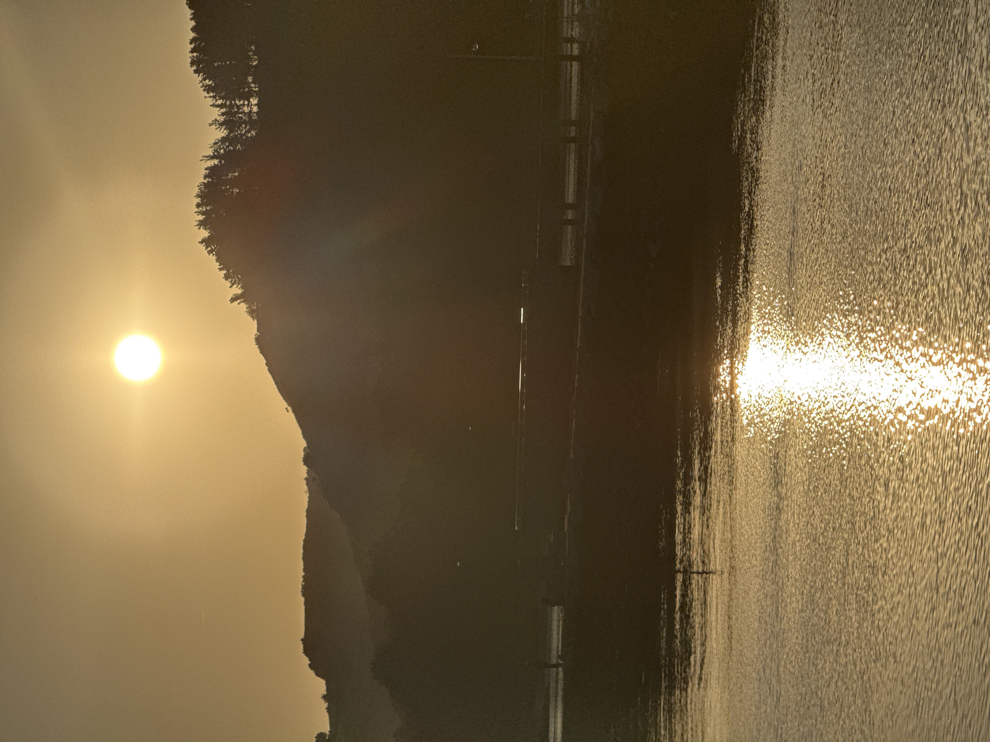 The image depicts a serene waterscape with a dramatic sunset over Zumaia, Spain. The sun, a bright orb, hangs in the hazy sky, casting a warm, golden glow over the scene. Silhouetted against the radiant sky are rolling hills covered in a dense forest of pine trees. In the middle ground, a bridge spans across a body of water, its structure partially obscured by the low light. A few indistinct figures are visible on or near the bridge, adding a sense of scale to the vastness of the landscape. The foreground is dominated by calm water, its surface shimmering with the reflection of the setting sun, creating a dazzling display of light and color. The overall atmosphere is one of tranquility and natural beauty, capturing a moment of quietude as day transitions into night.