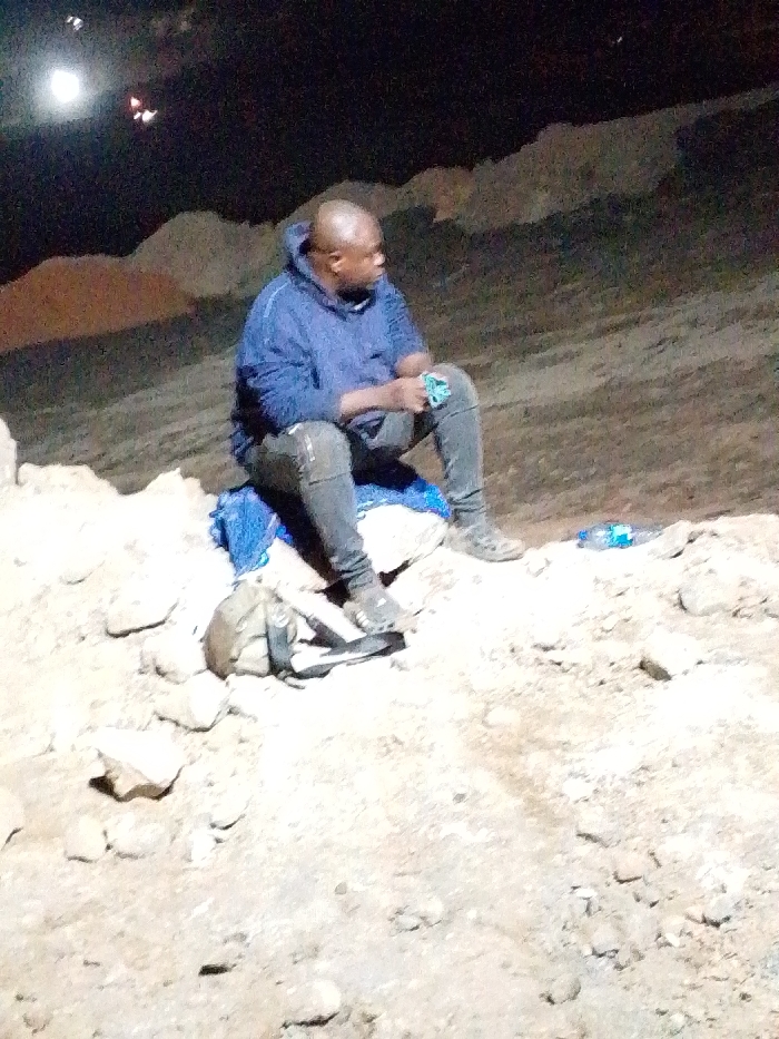 A person in a blue hoodie and gray pants sits on a rock formation in a dimly lit environment. The individual is looking to the right, holding something in their hands. A blue cloth and a backpack are near them on the rock. A water bottle is visible nearby. The surrounding area appears rocky and uneven.
