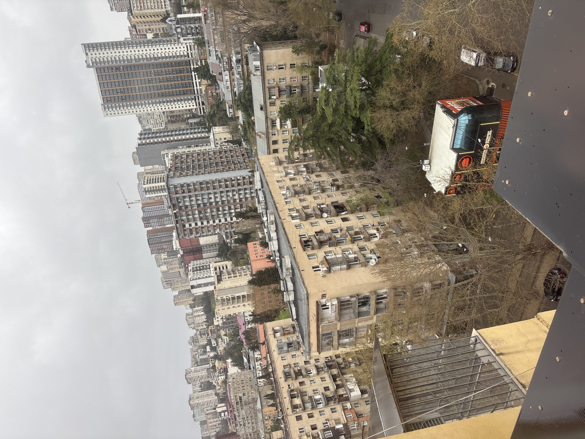 The image depicts an urban landscape in Tbilisi, Georgia, viewed from an elevated perspective. The scene is dominated by numerous high-rise and mid-rise buildings, densely packed and ranging in architectural styles. Many buildings are beige or light-colored with rows of windows and air conditioning units. A red double-decker tourist bus is visible, parked near a building. A few trees are scattered throughout the scene, some bare and others with dense green foliage. Several cars are parked along the street. The weather appears overcast, with a grey sky. The viewpoint seems to be from a high building with a metal structure visible in the foreground.
