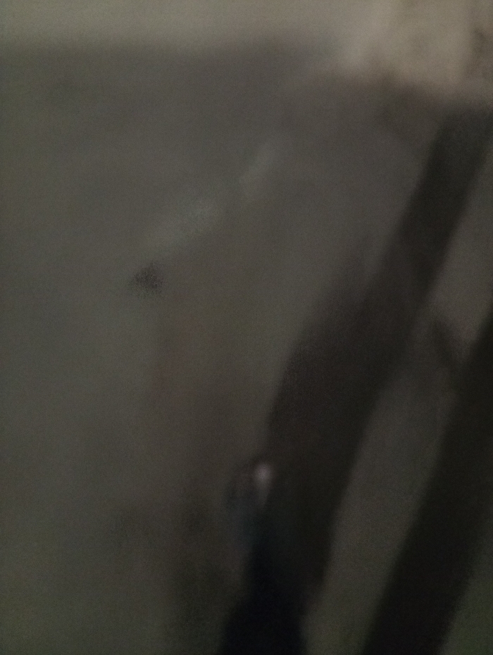 The image presents a dimly lit, mostly gray scene. Two dark, blurred vertical lines are visible on the right side, potentially shadows. A smaller, blurry dark spot appears on the upper left, accompanied by indistinct, faded marks around it. The overall impression is of a dark, indistinct environment with unclear shapes and forms.
