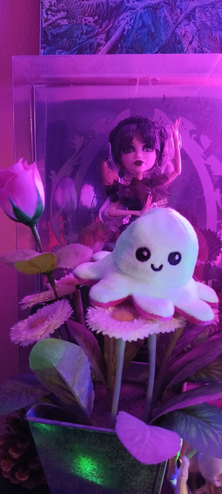 The image shows a close-up of a doll and a plush toy in front of a decorative background. The doll, dressed in dark attire with ruffled accents, has dark hair styled in braids and a pale complexion. Beside it, a light-colored plush octopus toy with a smiling face and wide eyes is perched on top of artificial flowers. The flowers are predominantly white and pink with yellow centers, and some green artificial leaves are also visible. The scene is illuminated by purple and pink lighting, casting colorful reflections on the metallic pot at the bottom of the image. The overall setting appears to be indoors, possibly a display or shelf. There is no visible indication of the city of Wels, Austria.
