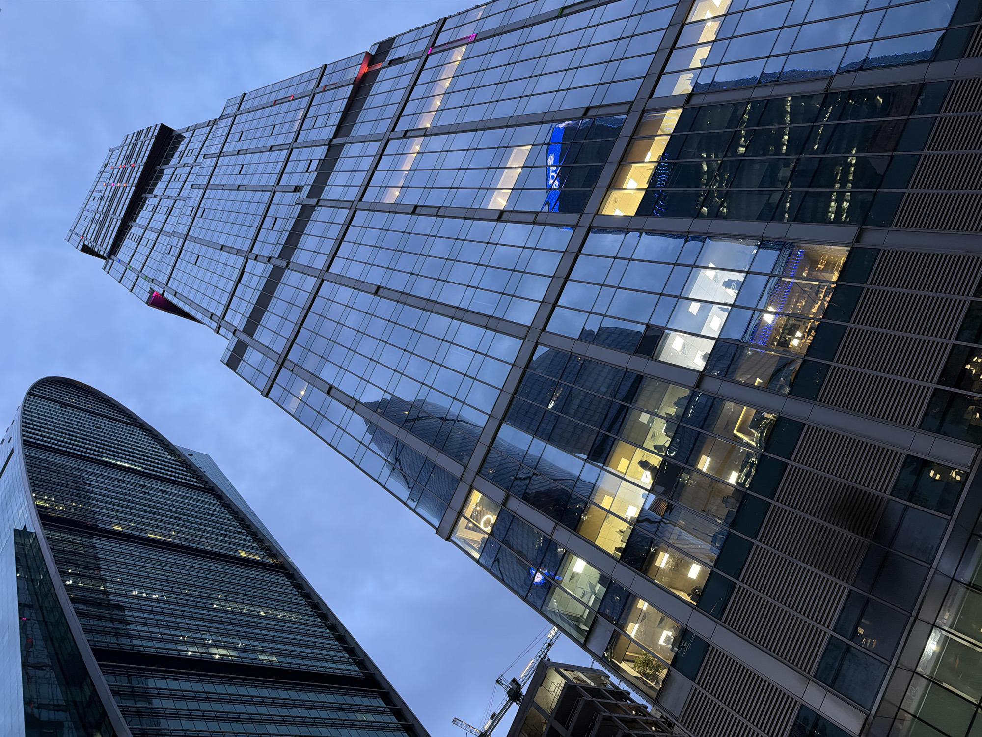 This media file depicts a low-angle, upward view of modern skyscrapers against a twilight sky. The main subject is the architecture of these tall buildings, characterized by their extensive glass facades that reflect the dimming sky and the illuminated interiors. One building on the left has a distinctive curved, segmented design, while the taller building in the center and right is more angular, featuring a stepped or tiered structure.
The reflections on the glass reveal glimpses of other buildings, interior office spaces with illuminated desks and chairs, and possibly structural elements of ongoing construction, indicated by a visible crane in the lower right portion of the image. The lights inside the buildings are a mix of warm yellow and cooler blue hues, creating a dynamic interplay of colors on the building surfaces. Small, colorful lights (red, pink, and blue) are also visible on the upper sections of the central building.
The time of day is clearly dusk or early evening, as suggested by the deep blue of the sky, the lack of direct sunlight, and the illuminated windows of the office buildings, implying that people are still working or that the lighting systems are on. The weather appears calm, with no visible signs of rain or strong winds.
While the location is specified as Moscow, Russia, there are no overt textual cues within the image itself (like signs or landmarks) to confirm this. The style of architecture, however, is consistent with modern urban centers. The overall mood is one of urban grandeur and activity, with a hint of introspection conveyed by the illuminated interiors seen through the glass. No people are explicitly visible, but their presence is implied by the lit offices.
