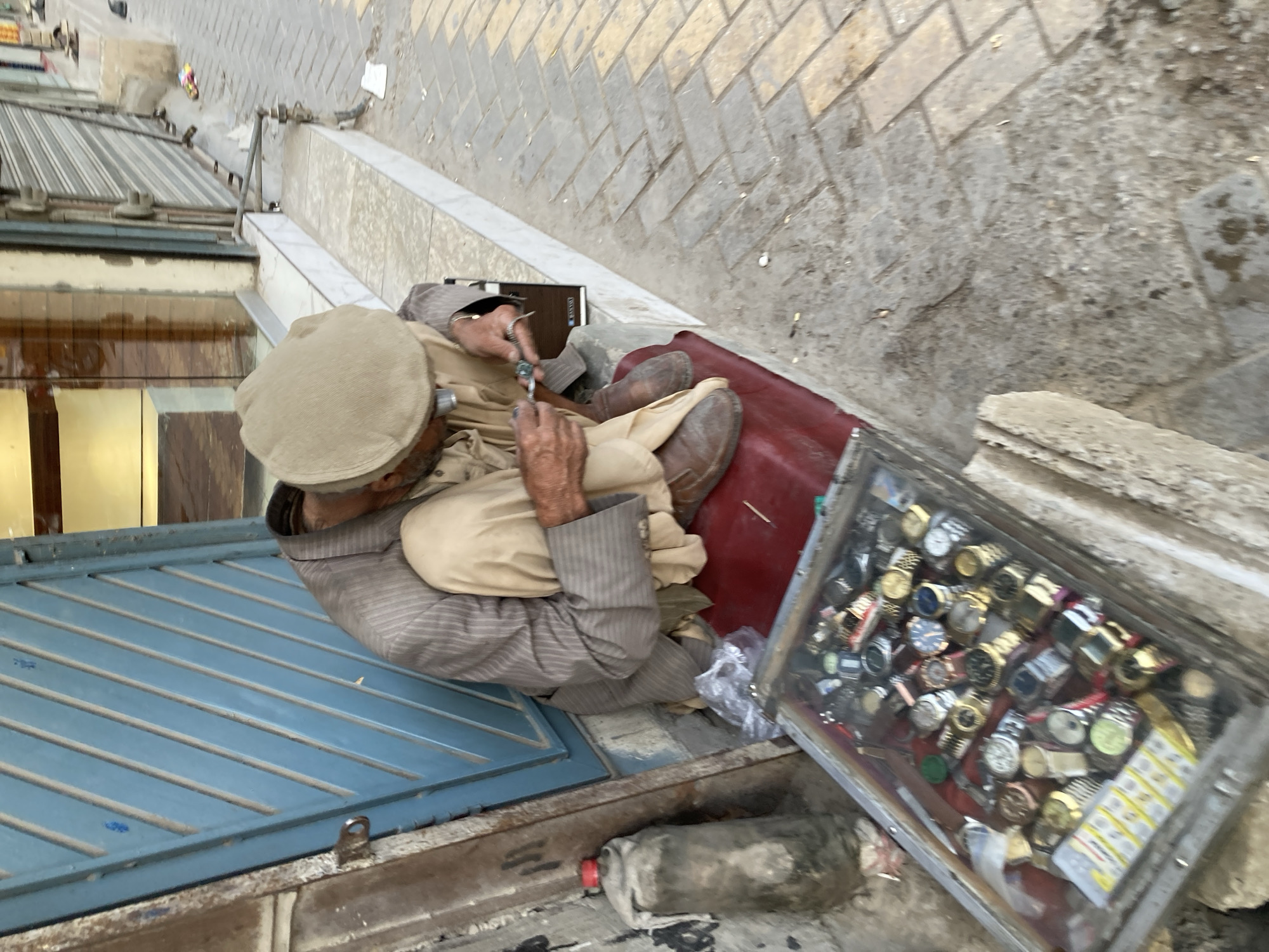 An overhead view captures an elderly man seated on the ground, seemingly engaged in work as a street vendor or repairer in an urban setting. He wears a light-colored cap, a striped shirt, and light-colored traditional trousers (likely shalwar kameez). His hands are focused on a small object, possibly a watch, holding a delicate tool, suggesting he is repairing or inspecting it.
To his right, a rectangular glass display case filled with numerous wristwatches of various designs, colors, and sizes is visible. Some watches appear to be gold-toned, others silver, with different strap materials. Next to the display case, a white/yellow packet with small, illegible text is present. A dark red mat or fabric is beneath him, and a dark-colored case or box rests beside him. A partially obscured plastic bottle with a red cap is also on the ground nearby.
The man is positioned next to a blue corrugated metal surface, which could be a closed shop shutter or part of his stall. The ground features light-colored paving stones and a white curb or step. The overall impression is of a bustling street scene, typical of Quetta City Tehsil, Pakistan, during daylight hours. No strong shadows indicate midday sun, suggesting morning or afternoon. The man appears focused on his task, contributing to a snapshot of daily life.