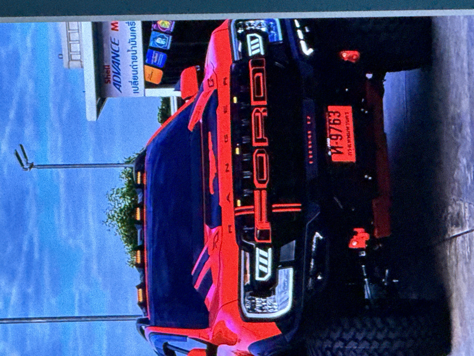 The image shows the front of a red Ford Raptor pickup truck parked outdoors. The truck has prominent "FORD" lettering across the grille, illuminated with red light. It also features unique headlights with white LED accents and additional orange lights on the roof. In the background, a building with "Shell ADVANCE M" signage is visible, suggesting it might be a gas station or service center. The sky is blue with some clouds, and a street light pole is present to the left. The license plate on the truck is red and appears to have Thai script. The overall scene is a bright, daytime shot focused on the vehicle. There are no people visible in the image. The location in the city of Cahul, Moldova is not discernible from the visual information present in the image.