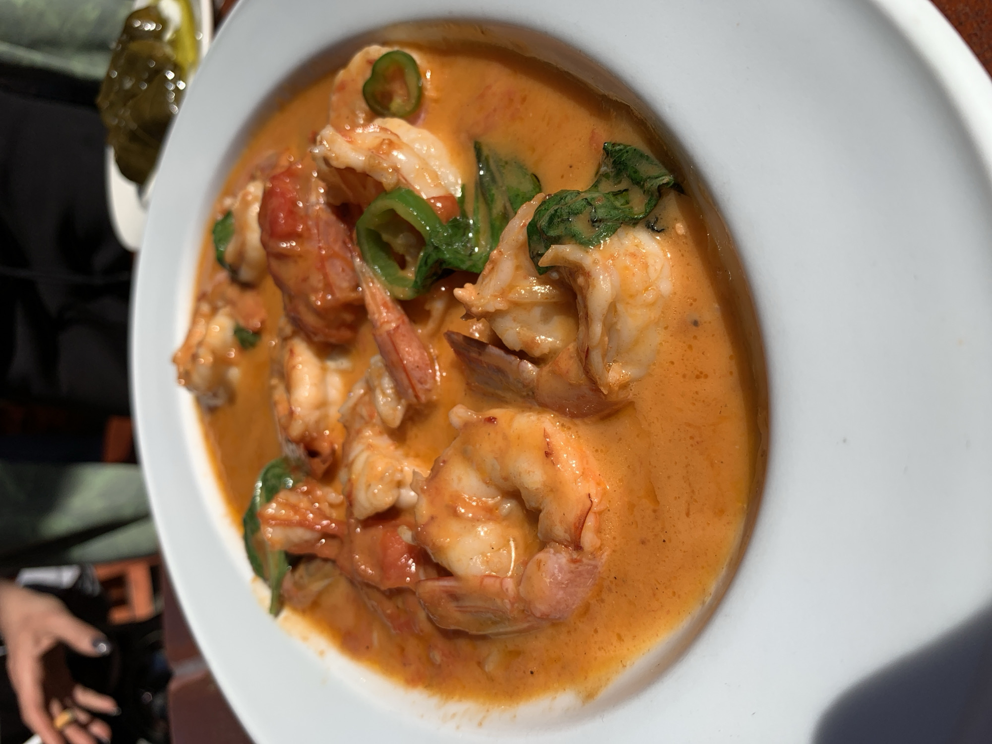 A close-up shot shows a white bowl filled with shrimp in an orange sauce, garnished with green chilies and spinach. The dish is likely part of a meal. In the background, a person's hand is visible, suggesting someone is either about to eat or has been eating the dish. The image was taken outdoors, under sunlight, likely in Tel-Aviv, Israel. A side dish of pickles is visible in the corner.