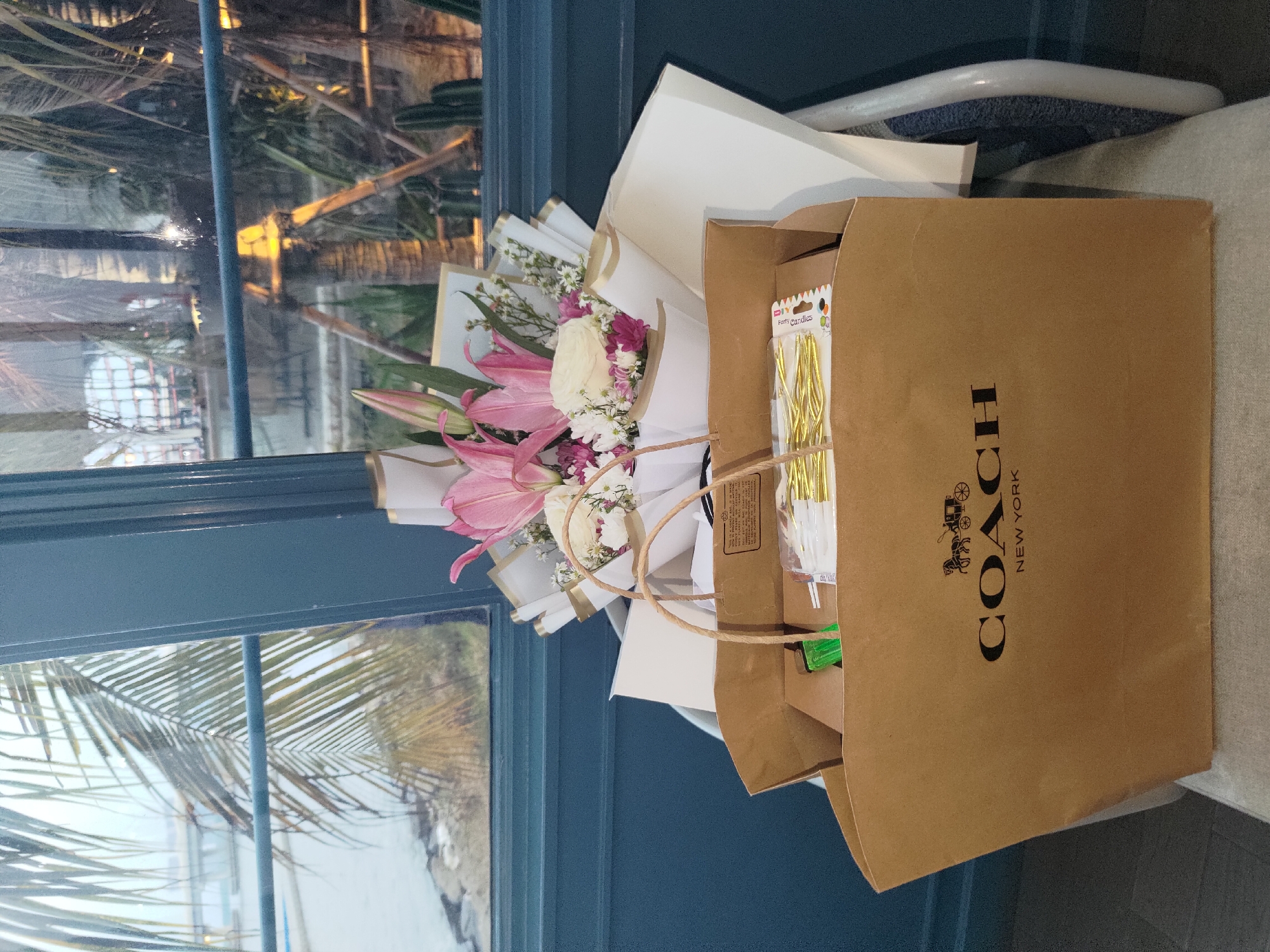 The image shows a Coach shopping bag with a bouquet of flowers on top, placed on a chair. The bag is brown with the Coach logo and "New York" printed in black. The bouquet consists of pink lilies and white roses, wrapped in white and gold paper. Inside the shopping bag, there are some party candles. In the background, through a window, a body of water, palm trees, and some buildings are visible, suggesting an outdoor or waterfront view. The scene appears to be indoors, possibly in a cafe or lounge, given the presence of furniture and the ambient lighting. The location is specified as Kosambi Barat, Indonesia. There are no people visible in the image.