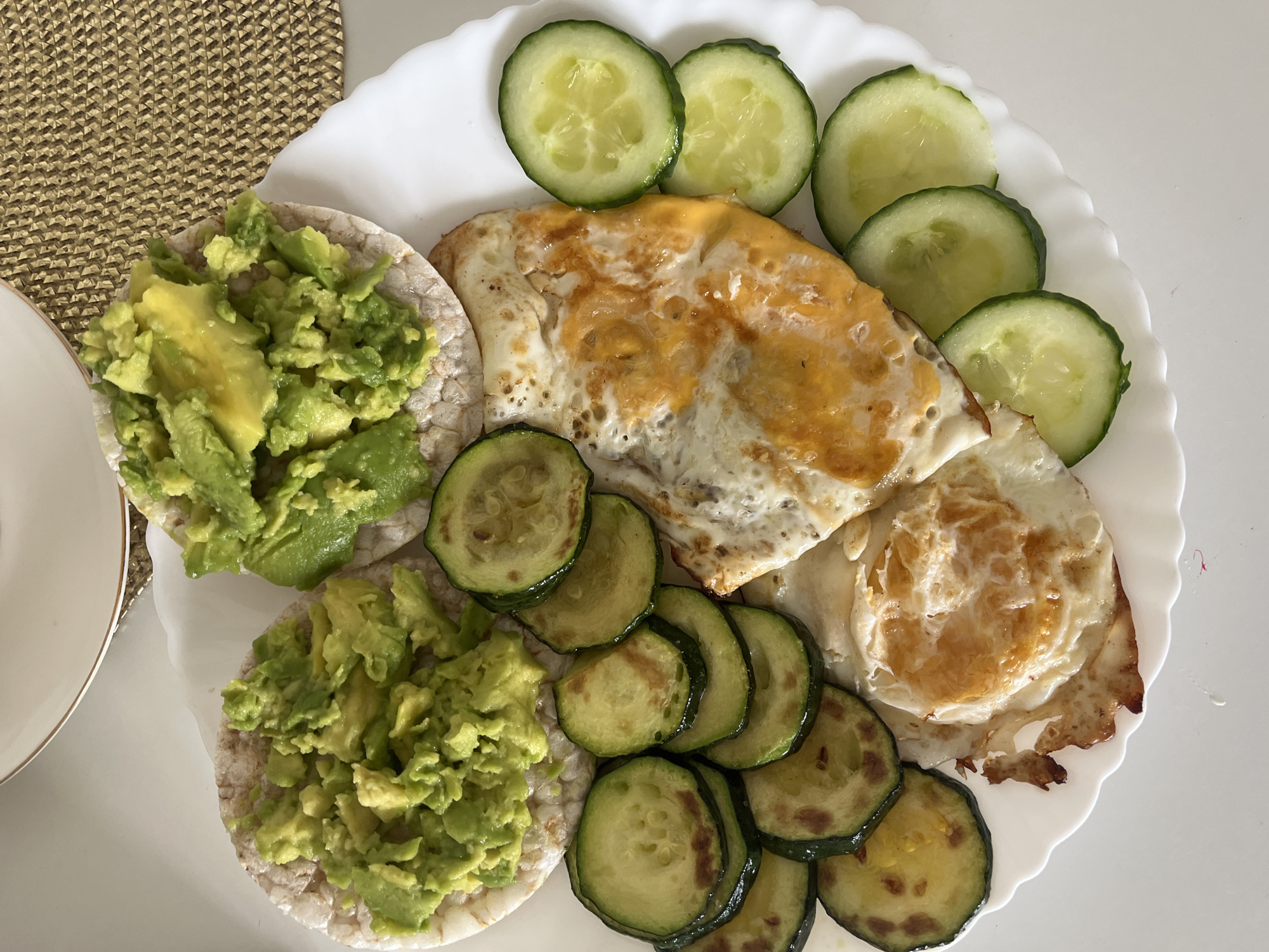 A plate of food is presented on a white surface, possibly a table, with a textured placemat visible in the upper left corner. The meal consists of several components arranged on a white, scalloped-edge plate. On the left side of the plate are two round rice cakes, each topped with a generous portion of mashed avocado. In the center and right of the plate, there are two fried eggs, one appearing to be a single fried egg and the other a partially cooked egg with melted cheese. Surrounding the eggs are several slices of cooked zucchini and a few slices of raw cucumber. The lighting suggests it is daytime. There is no visible text in the image.