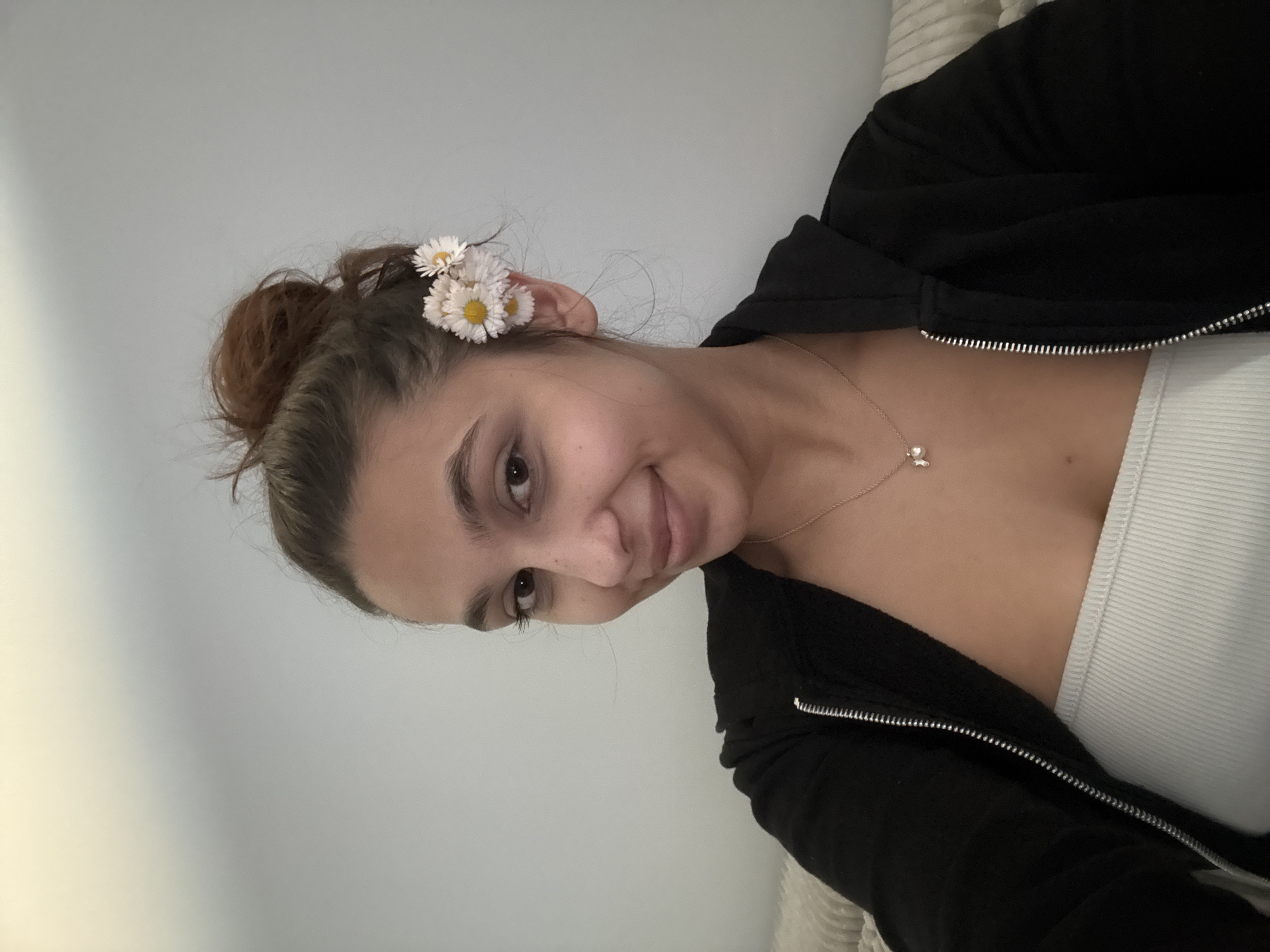 A young woman is photographed indoors. She has light skin, brown eyes, and dark eyebrows. Her brown hair is styled in a messy bun with three small white daisies tucked behind her right ear. She wears a white tank top under a black zip-up hoodie. A delicate gold necklace with a small pendant is visible around her neck. The background is a plain light gray wall. No specific details indicate the location is in Mönchengladbach, Germany.