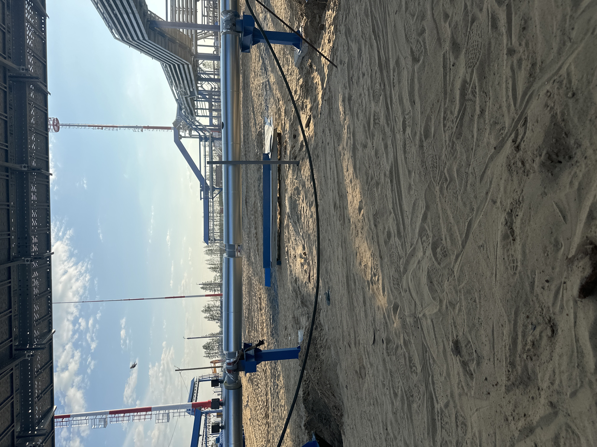 The image depicts an industrial construction site under a clear blue sky with scattered clouds. In the foreground, a sandy ground is covered with numerous footprints, suggesting recent activity. A large, silver-colored pipe runs horizontally across the mid-ground, supported by blue metal structures. To the left, a red and white striped tower is visible, and further back, more industrial infrastructure, including elevated pipelines and other towers, can be seen. The environment appears to be an open area with a few distant trees on the horizon. The overall scene conveys a sense of ongoing industrial development in a relatively undeveloped area, consistent with the location in Chikshino, Russia, which is known for its oil and gas infrastructure development.