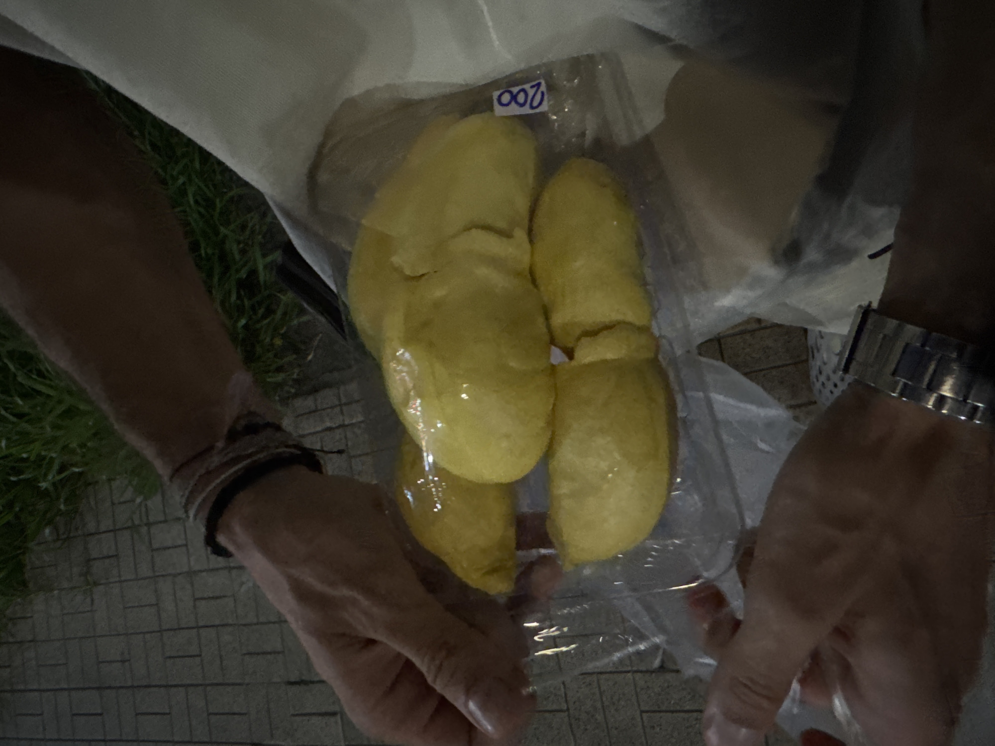 In an outdoor setting with a cobblestone walkway and grass in the background, a person is holding a clear plastic bag containing several pieces of yellow durian. The bag is sealed and has a blue sticker with the number "200" written on it. The person's hands are visible, with one hand holding the bag from the top and the other supporting it from the bottom. The person is wearing bracelets on their left wrist and a watch on their right wrist. The lighting suggests it is evening or night. The location is identified as Surat Thani City Municipality, Thailand.