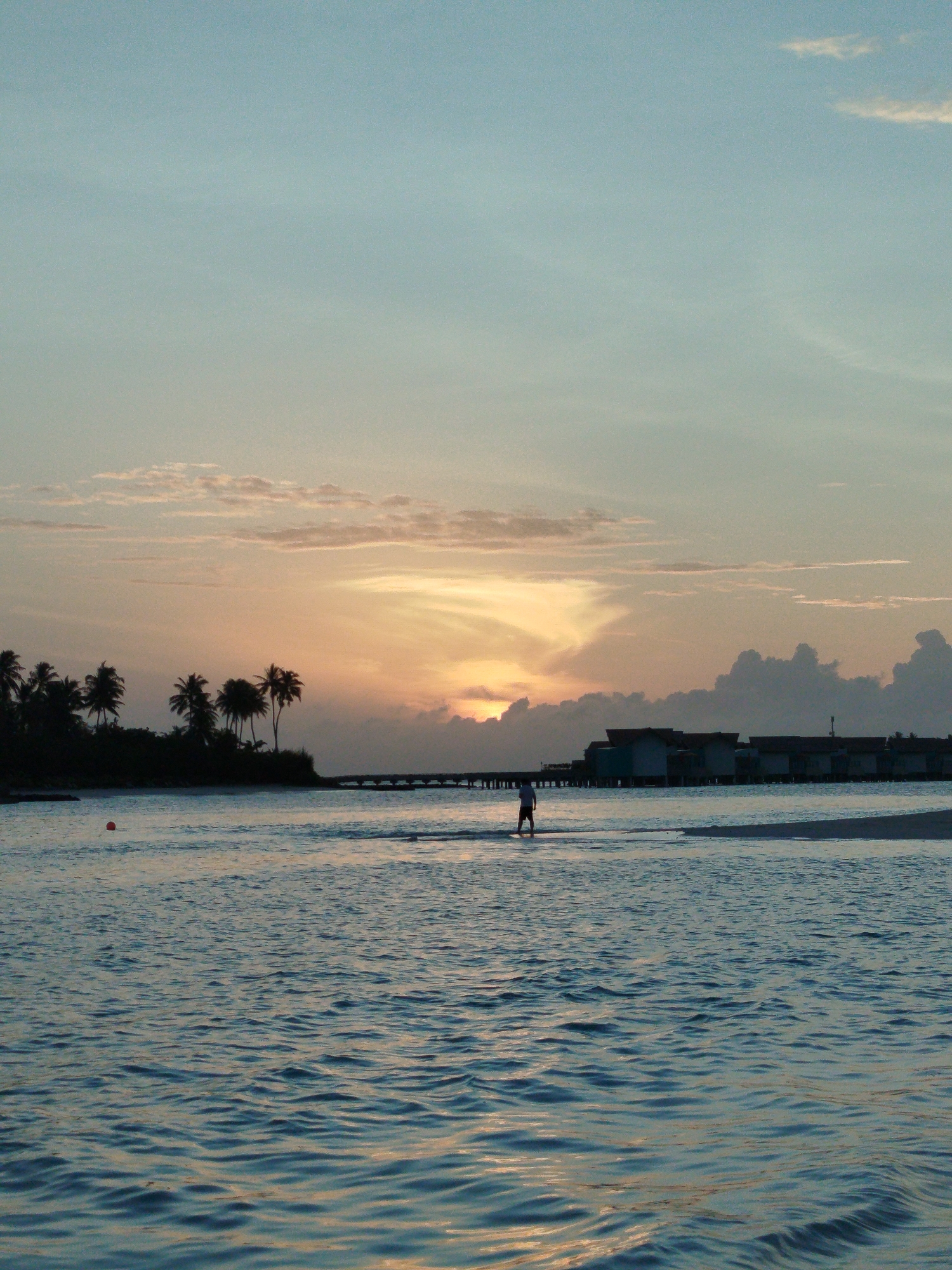 The image shows a serene sunset scene in the Maldives. The sky is a gradient of soft blues and oranges, with scattered clouds illuminated by the setting sun. In the foreground, calm ocean waters reflect the sky's colors, with gentle ripples on the surface. A lone figure, silhouetted against the bright sky, stands in the shallow water. To the left, a line of palm trees is visible against the horizon, suggesting a tropical island. In the distance, a pier extends into the water, and beyond that, several overwater bungalows are clustered together, characteristic of Maldivian resorts. The scene evokes a sense of tranquility and natural beauty, typical of a sunset on a tropical island.