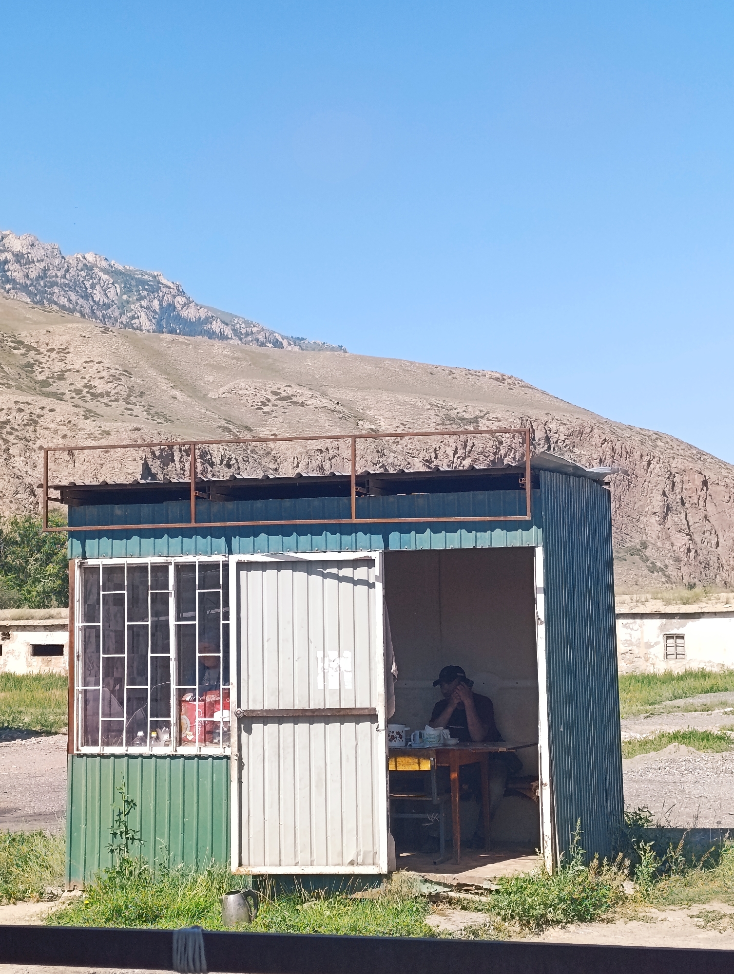 The image depicts a small, corrugated metal kiosk or guardhouse situated in a semi-arid, mountainous environment, likely within the city of Naryn, Kyrgyzstan. The structure is painted in sections of green and blue, with a corrugated metal roof covered by a rusted metal frame. A person wearing a dark shirt and a baseball cap is seated inside the kiosk at a table, appearing to be looking down. On the table are several mugs, plates, and possibly a kettle. To the left of the kiosk, a window with white bars is visible, showing some items inside, including what appears to be a red can. In the background, a large, barren mountain range dominates the skyline under a clear blue sky. The ground around the kiosk is unpaved, with some sparse green vegetation growing in patches. To the right of the kiosk, another smaller building with windows is visible. The overall scene suggests a remote or functional outpost in a natural setting.