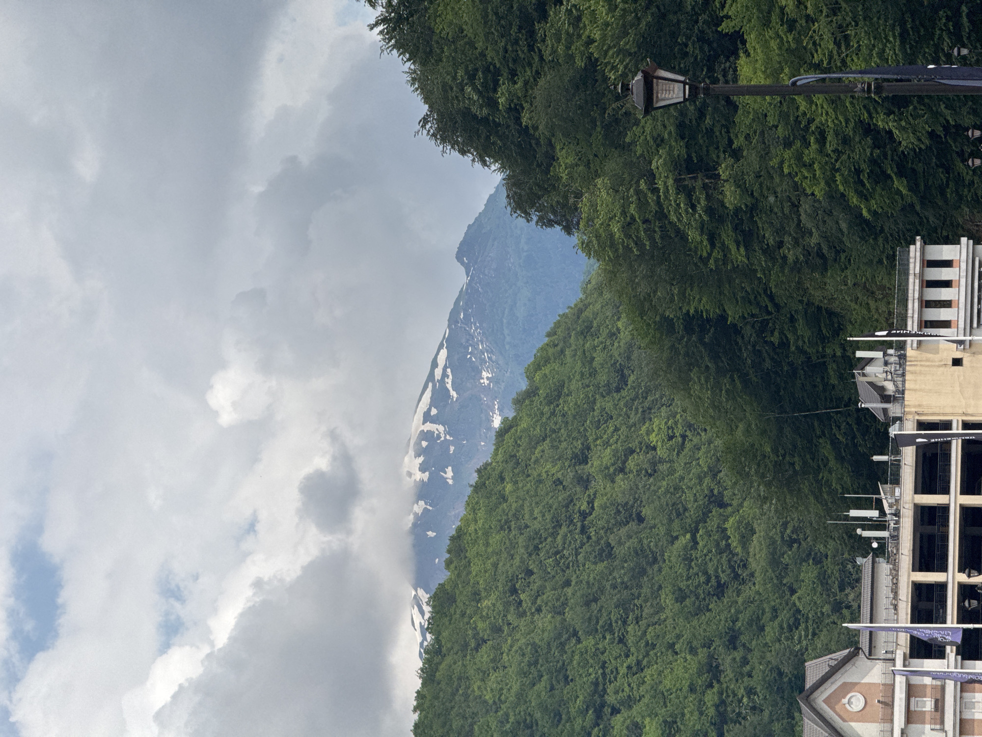 A landscape scene in Estosadok, Russia, features a snow-capped mountain peak partially obscured by clouds in the background. The mountain is surrounded by lush green, forested slopes. In the foreground, a multi-story building with an open-air lower level and a distinctive tiled roof is visible. A lamppost stands to the right of the building, and flags are displayed on poles. The sky is overcast with patches of blue.