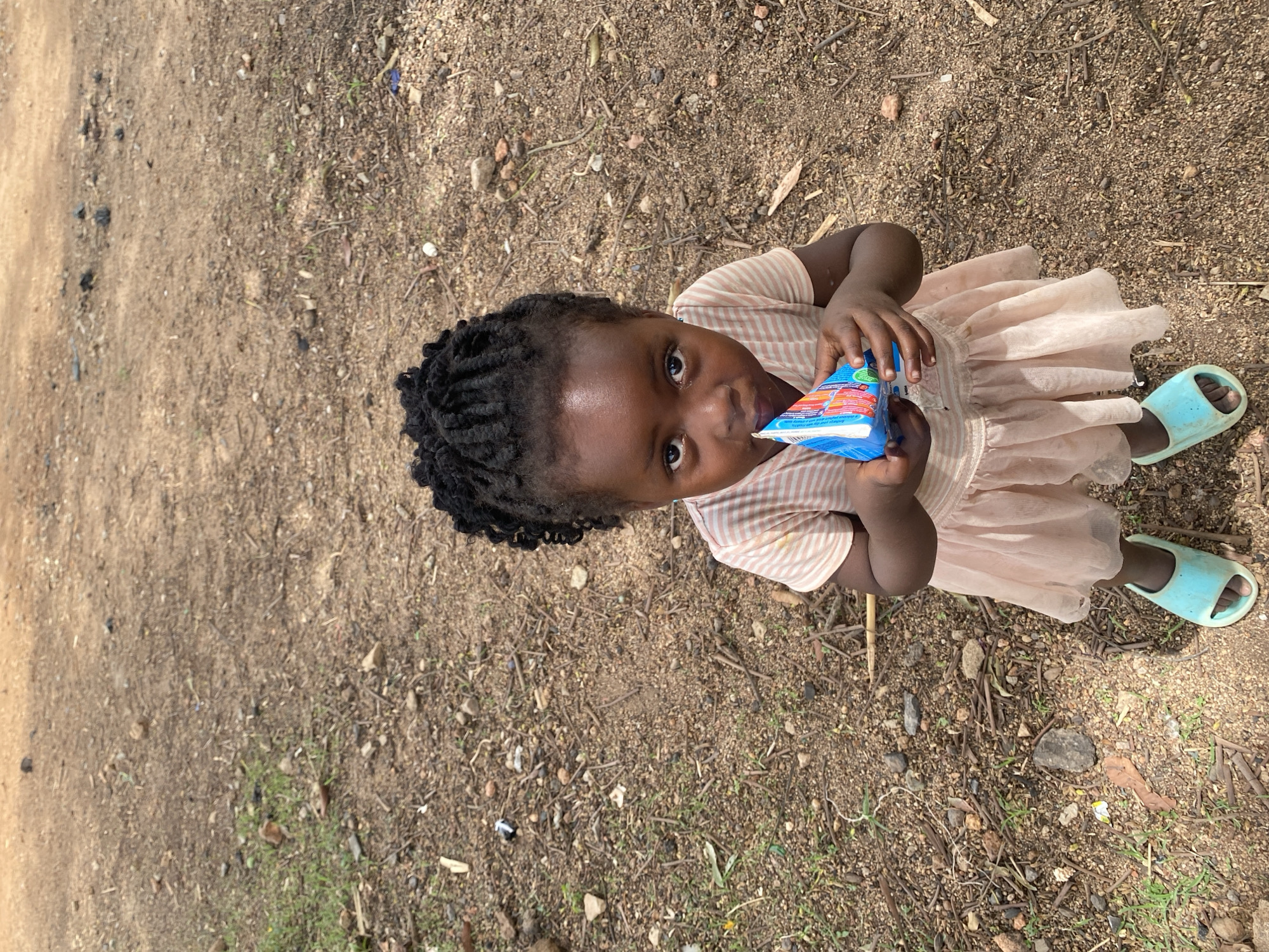A young, dark-skinned child with braided hair is standing on a dirt and gravel surface, presumably in Kuru Dungus, Nigeria. The child is holding a blue and red juice box and drinking from it. The child wears a pink and white striped shirt with a light pink tulle skirt and blue sandals. The ground is mixed with loose dirt, small rocks, and scattered dry twigs. Patches of sparse green grass are visible. A dirt path is visible in the upper left.