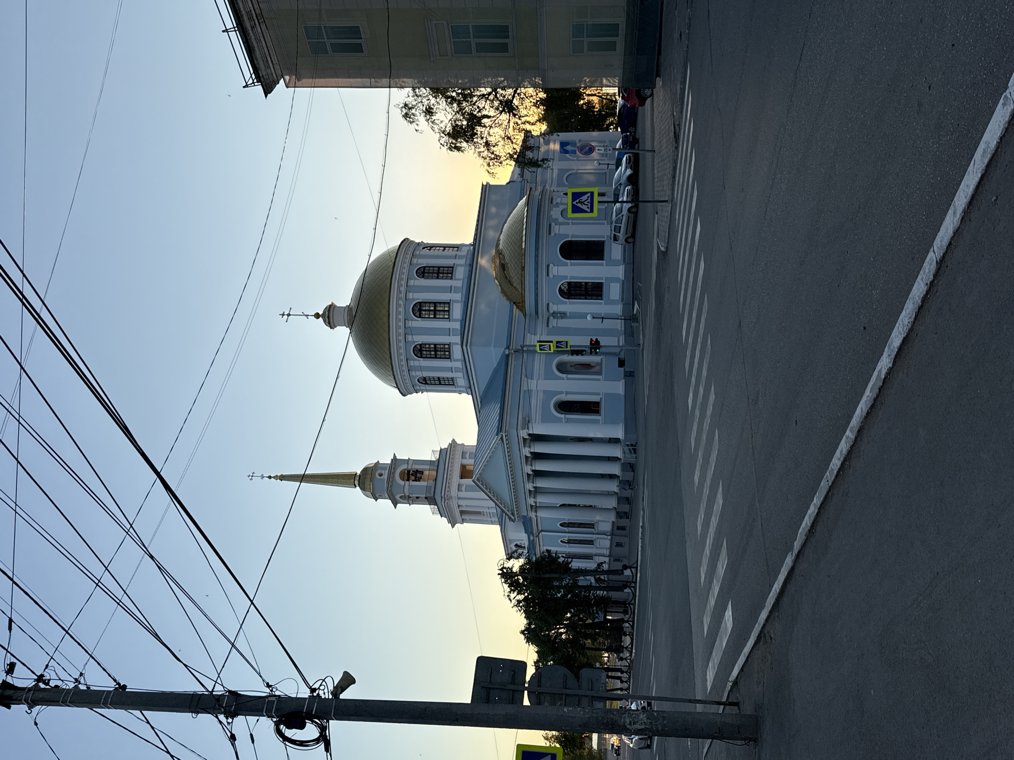 The image depicts the Church of the Intercession of the Holy Virgin in Votkinsk, Russia, at dusk. The church, a prominent blue building with a golden dome and bell tower, is illuminated by the setting sun. A street with a pedestrian crossing is in the foreground, with a utility pole and traffic lights visible. Cars are parked near the church, and several trees are scattered around the area. The sky is clear and light blue, transitioning to a pale yellow near the horizon. The overall scene is serene and picturesque, capturing the beauty of the church and its surroundings during the golden hour.