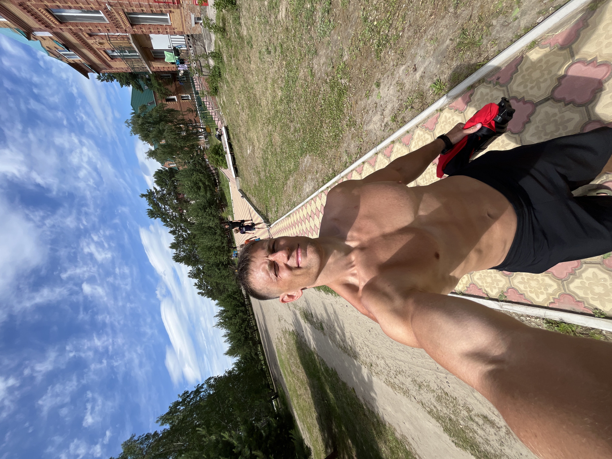 In Lebyazhye, Russia, a shirtless man takes a selfie outdoors on a sunny day. He is wearing black shorts and holding a red and black item in his right hand. The man is standing on a patterned tiled pathway. Behind him, a dirt path runs parallel, bordered by grass and trees. In the background, there are several brick buildings with green roofs and a clear blue sky with scattered white clouds. Other people are visible in the distance, walking along the paths and near the buildings.