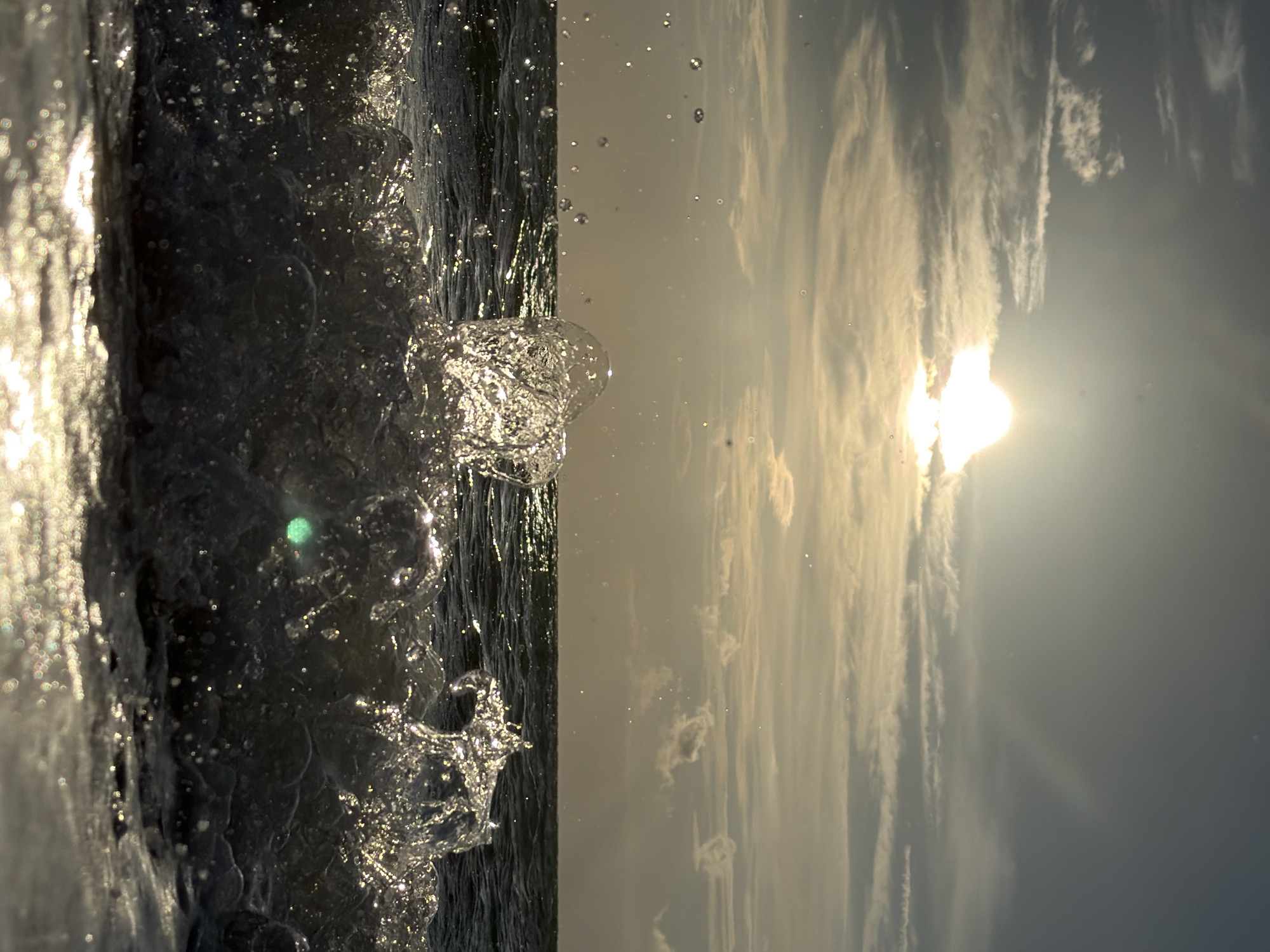 A close-up, low-angle shot captures a crashing wave against a beach. The sun shines brightly in the upper portion of the frame, partially obscured by clouds and casting a golden hue over the scene. The water is dark and appears choppy, with splashes and spray frozen in motion. The foreground is dominated by the crest of the wave, a dynamic explosion of water droplets, with sunlight glinting off its surface. The indistinct horizon suggests an ocean or sea. The location is not specified in the image.