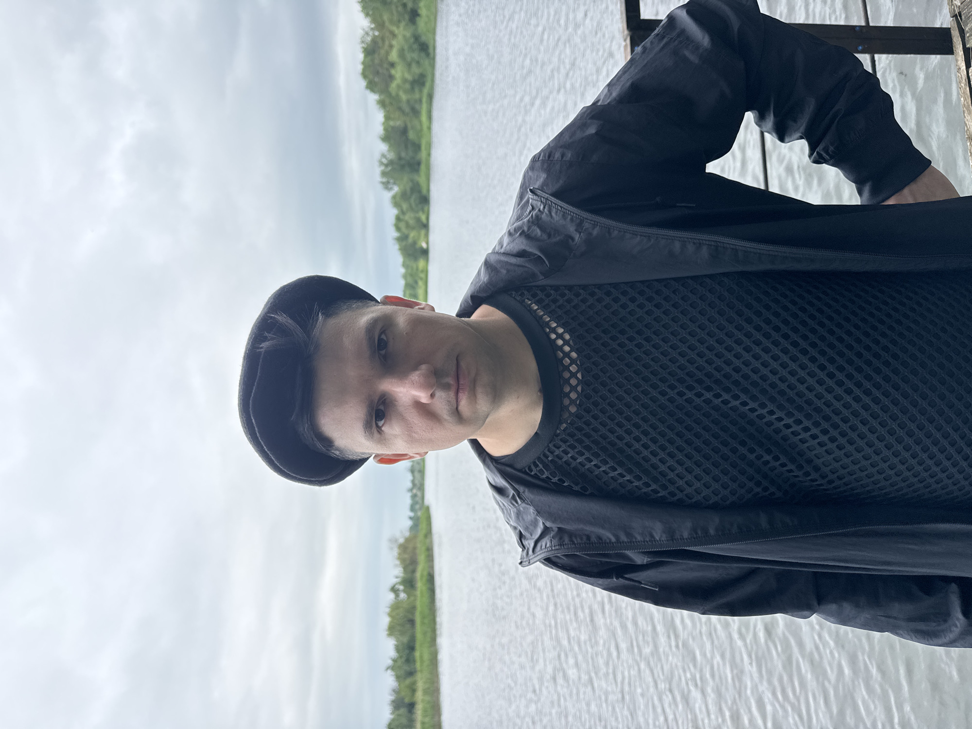 A medium shot captures a man wearing a black beret, a black mesh long-sleeved shirt, and a black zip-up jacket with his right hand in his pocket. He stands outdoors with a body of water behind him, and a tree line in the distance. The sky is overcast. The location is identified as Lozuvatka, Ukraine.