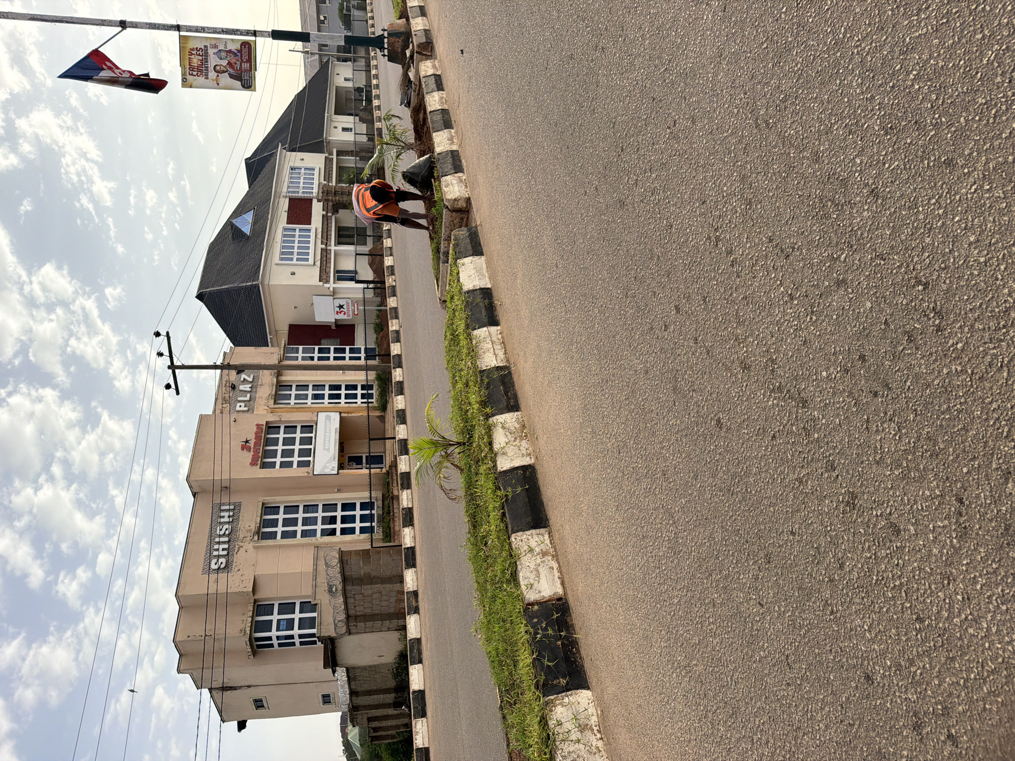 The image shows a street scene in Bukuru, Nigeria. In the background, there are two buildings. The left building, painted a light tan, has the word "SHISH!" in large letters above several windows. To its right is another building with the word "PLAZ" visible, and a sign for "3* SUPERMARKET". The right building is a lighter color with a dark, gabled roof. A flag is visible on a pole to the right of the buildings, and below it is a banner with images of two men. In the foreground, a person wearing an orange vest and dark shorts is tending to a small plant in a median strip. The median strip is lined with black and white painted curbs and has grass growing in it. The street in front of the median is paved and appears to be asphalt. Overhead, power lines are strung across the sky, and the sky itself is partly cloudy.