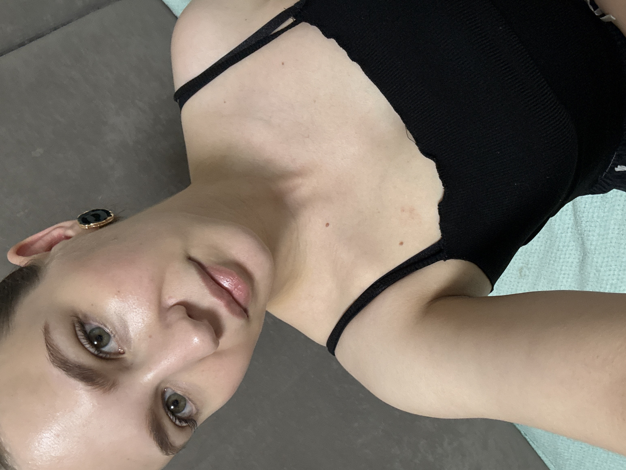 A young woman is shown in a close-up selfie. She has fair skin, green eyes, and dark eyebrows. She is wearing a black tank top and a black and gold earring in her right ear. Her head is tilted, and she is looking at the camera. The background is a gray textured surface, possibly a wall or furniture. The scene does not provide any specific location details within Chaikovsky, Russia.