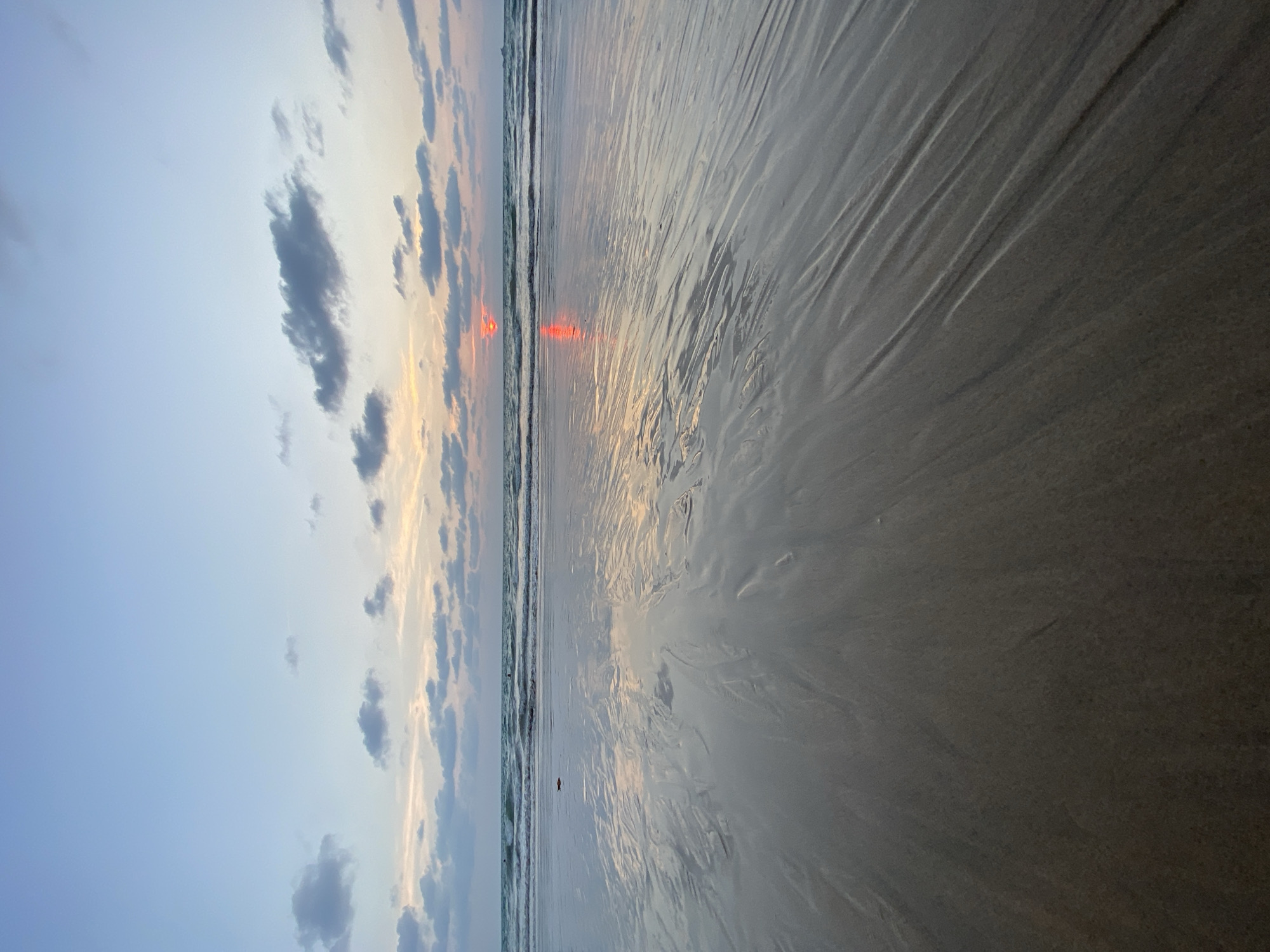 The image depicts a beach at sunset, likely in Choeng Thale, Thailand. The sky is a gradient of blue and orange, with scattered clouds. The sun's reflection stretches across the wet sand. The tide is low, revealing the sand's textures. There are several waves breaking on the shore.