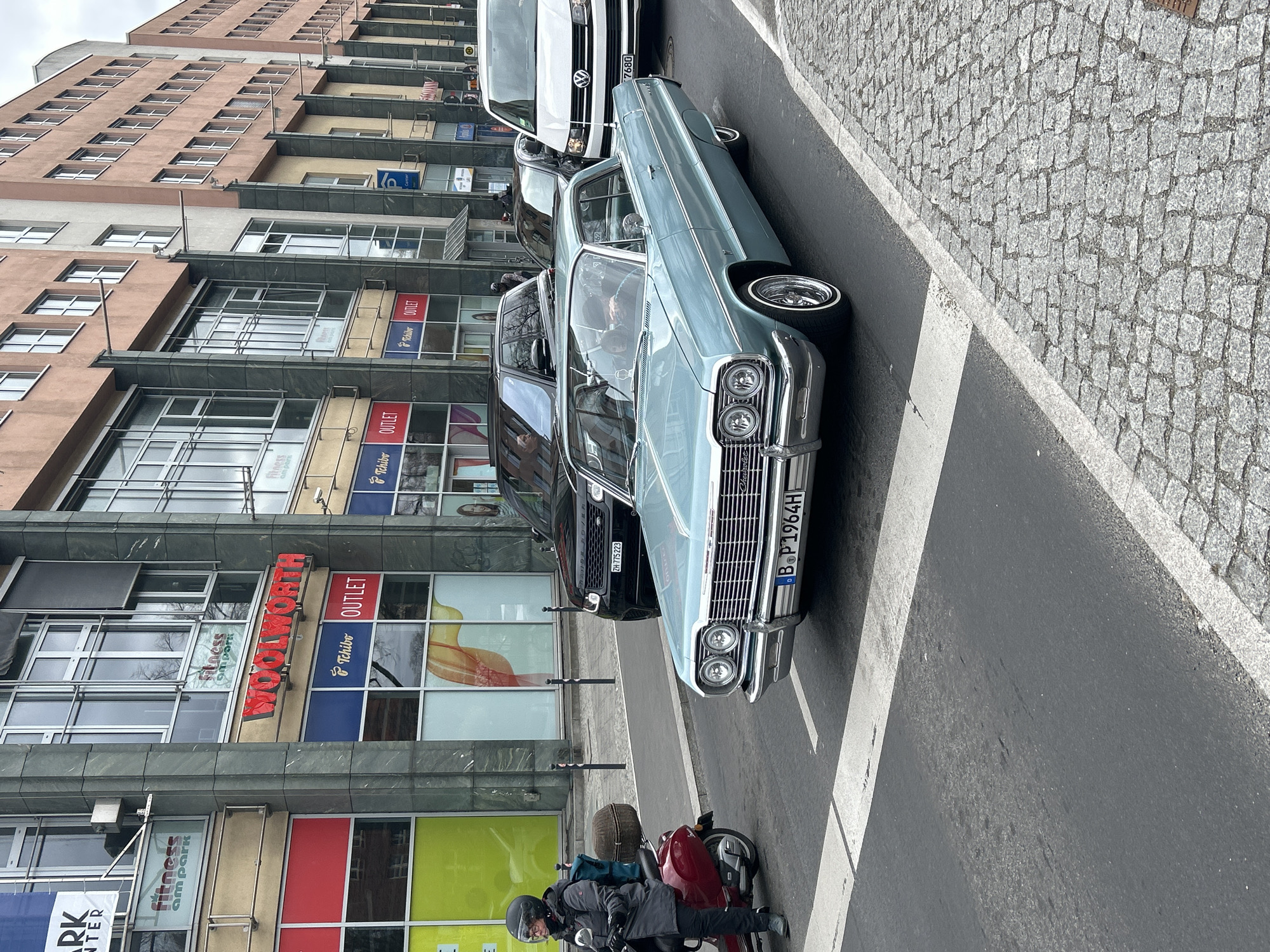 The photograph captures a street scene in Berlin, Germany, showcasing a vintage light blue Chevrolet Impala with the license plate "B·P1964H" parked on the street. Behind the Impala, there is a black Range Rover with the license plate "ZH-775223," followed by a white Volkswagen van with the license plate "7680." A person on a maroon motorcycle with a suitcase strapped to the back is positioned in front of the Impala, waiting or pausing on the side of the road. The building in the background features retail outlets, including "Fitness am Park," "WOOLWORTH," and "Tchibo OUTLET." The street consists of asphalt, with a crosswalk marking the road, while the sidewalk on the right is paved with cobblestones. The architecture and signage suggest a typical urban environment, and the variety of vehicles indicates a mix of contemporary and vintage automotive culture.