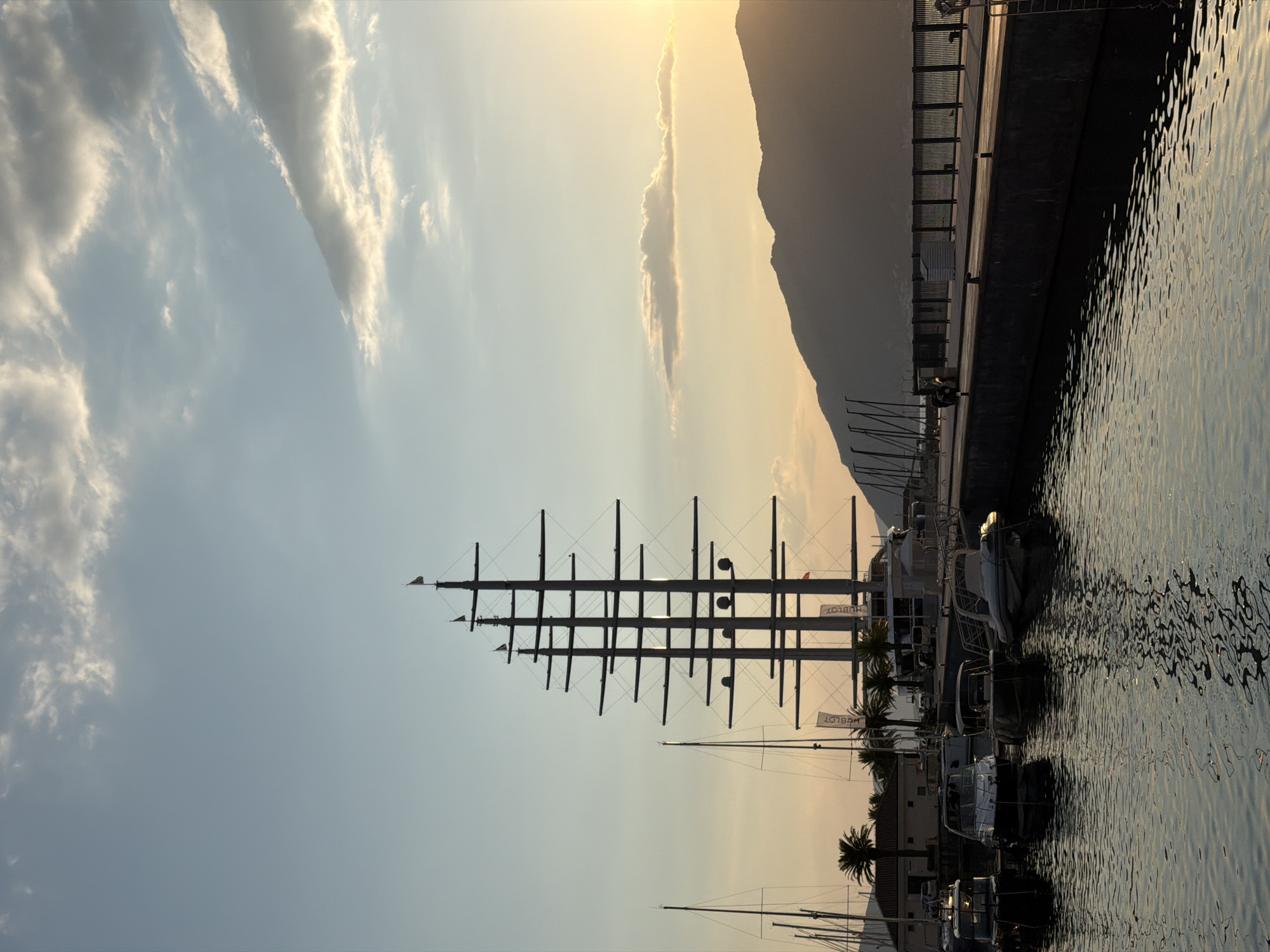 This image depicts a marina in Tivat, Montenegro, at sunset. The scene is dominated by the tall masts of several sailboats, silhouetted against a warm, golden sky. The sun is setting behind a mountain range, casting a soft glow on the clouds and the water. The water reflects the sky and the boats, creating a shimmering effect.
In the foreground, several yachts are docked, their sleek designs visible. Palm trees add a tropical touch to the scene. A long, concrete pier runs along the edge of the marina, with a fence bordering it. The overall mood is peaceful and serene, capturing the beauty of a Mediterranean evening.
Visible text: "HUBLOT" is printed on two flags hanging from poles near the sailboats.