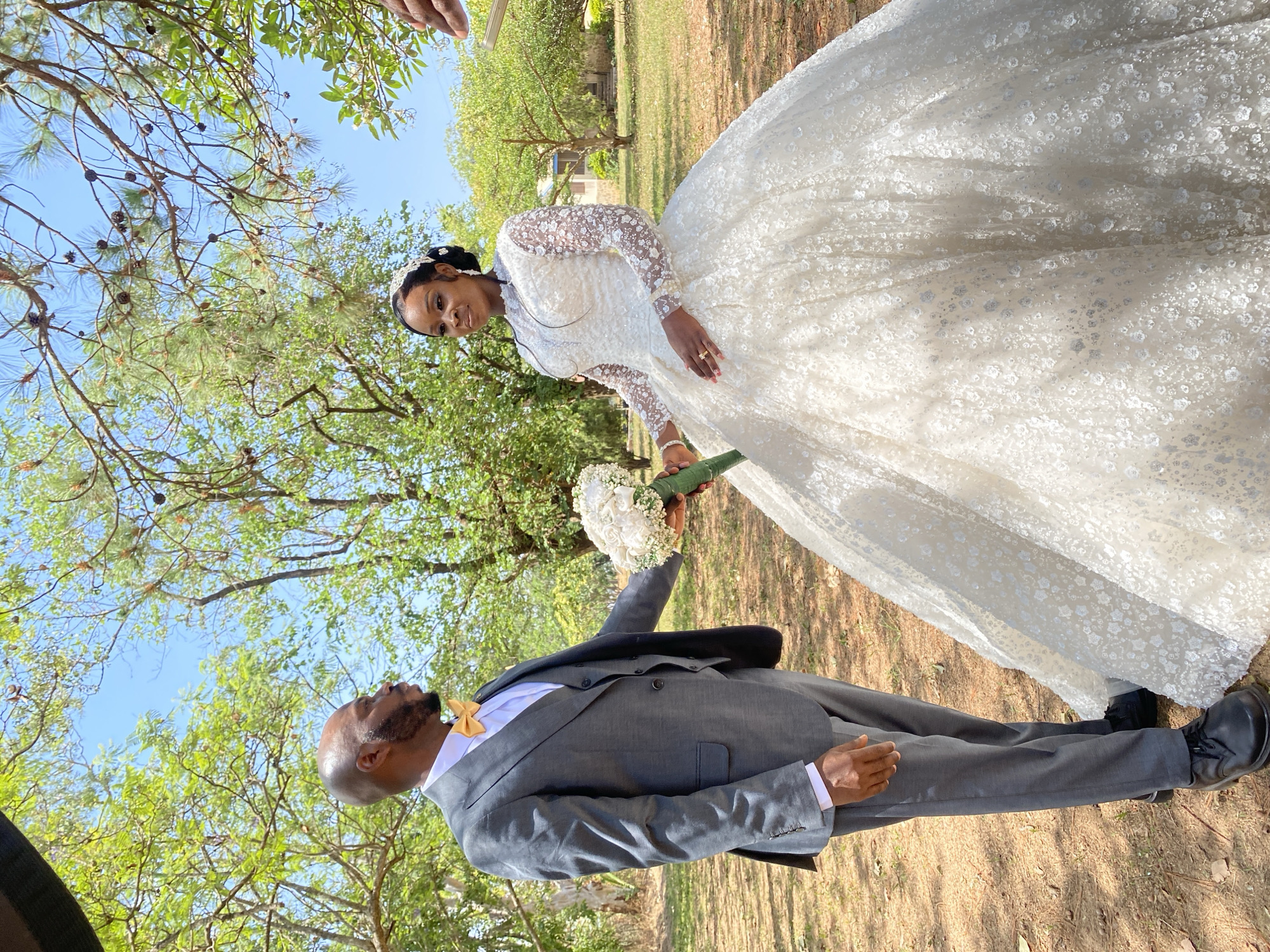 The photograph captures a wedding scene in Dutsen Kato, Nigeria. A Black bride in an elaborate white lace gown stands with a bouquet of white flowers. A Black groom in a grey suit with a gold bow tie stands opposite her, reaching for the bouquet. The background features trees and a clear blue sky, suggesting an outdoor setting.