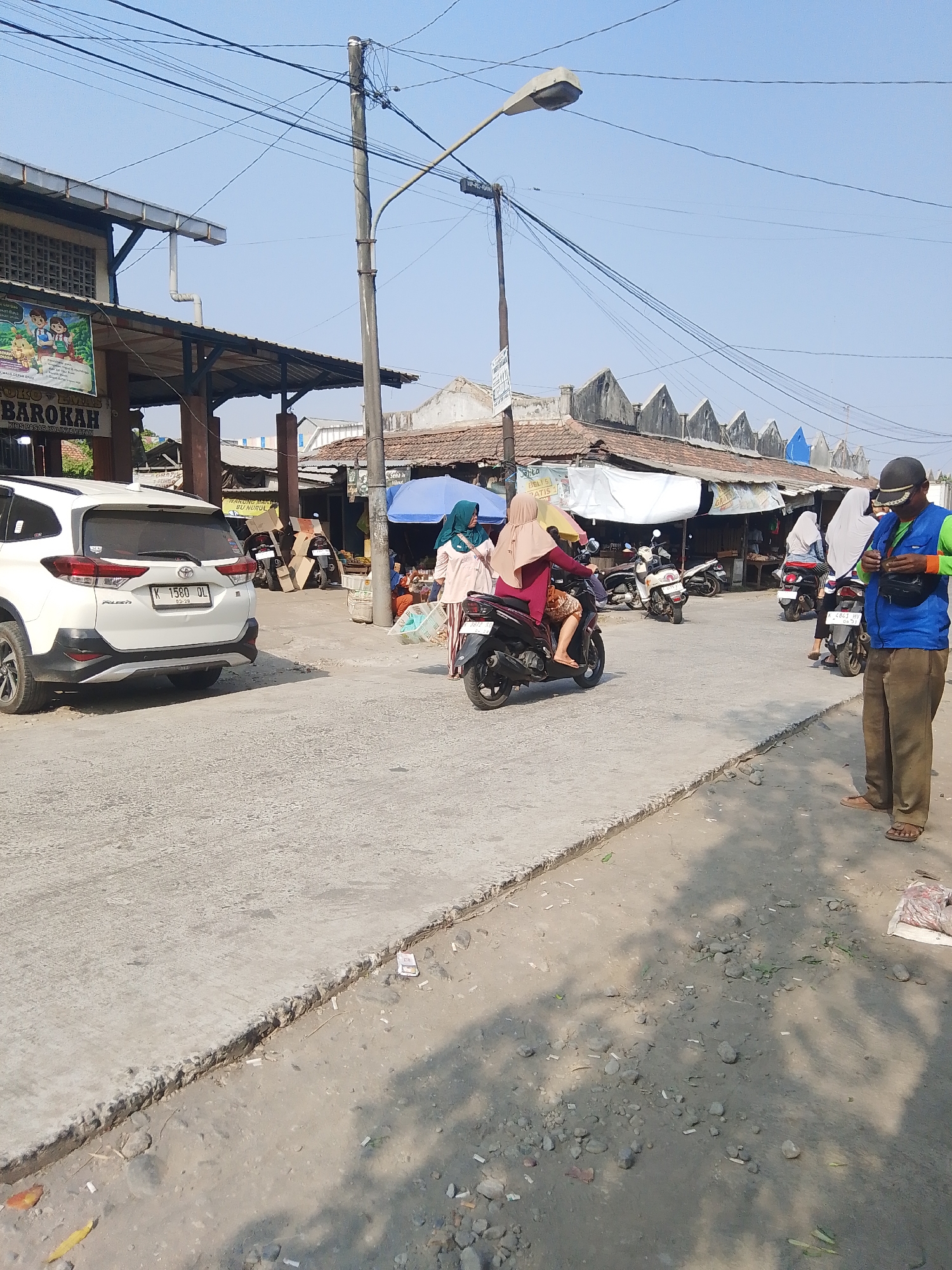 The image depicts a street scene in the city of Kalinyamatan, Indonesia. A white SUV is parked on the left side of a paved road, facing away from the camera. Beyond the car, market stalls are visible with awnings and various goods. Several people are present in the scene. One man, wearing a blue vest and brown pants, stands on the right side of the road, facing away from the camera and looking down at something he is holding. Several individuals are riding motorcycles, with one woman in a maroon hijab and dress on a scooter in the center of the frame. Another woman in a light blue hijab and a light-colored top is walking on the right side of the road. The environment appears to be a sunny day, with clear blue skies. The road is a mix of paved and dirt sections, with debris and cigarette butts scattered on the ground. Overhead, a complex network of electrical wires is visible, strung between poles. The architecture of the market stalls features tiled roofs and wooden supports.