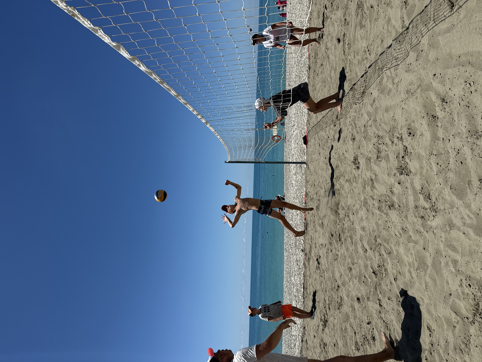 This is a ground-level, outdoor, full shot of a beach volleyball game on a sunny day in Nebug, Russia. The scene depicts a volleyball net set up on a sandy beach with the Black Sea and a clear blue sky in the background. There are at least five people visible playing volleyball. One individual, a young male, is in mid-air, jumping to hit the ball, which is also in the air. Another player, a young female wearing a white cap, is positioned to receive or hit the ball on the other side of the net. Two other players are visible on the left side of the net, one adult male and one young male, and another player is partially visible on the right side. The sand is light brown, and the water is a deep turquoise. The overall impression is of a recreational activity taking place on a warm day.