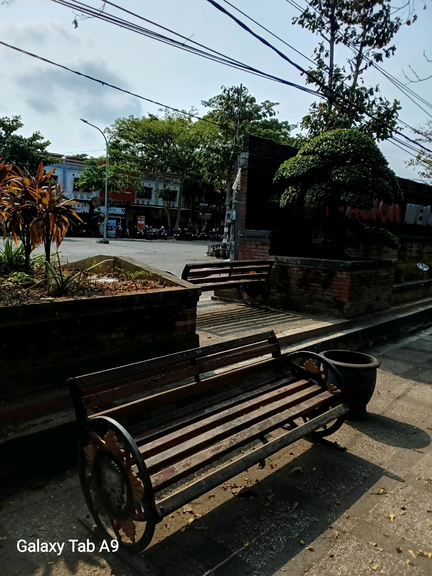 The image depicts an outdoor scene, likely a public area, with two wooden benches in the foreground and middle ground. The bench in the foreground is made of dark wood slats with decorative metal supports. A large dark pot sits to the right of this bench. Behind the foreground bench, another bench is visible, made of similar materials. A concrete planter with dry vegetation sits to the left of this second bench. In the background, a street is visible with buildings and parked motorcycles. Trees and power lines are present throughout the scene. The overall environment appears to be a sunny day with shadows cast on the ground. The image contains a watermark in the lower left corner reading "Galaxy Tab A9".