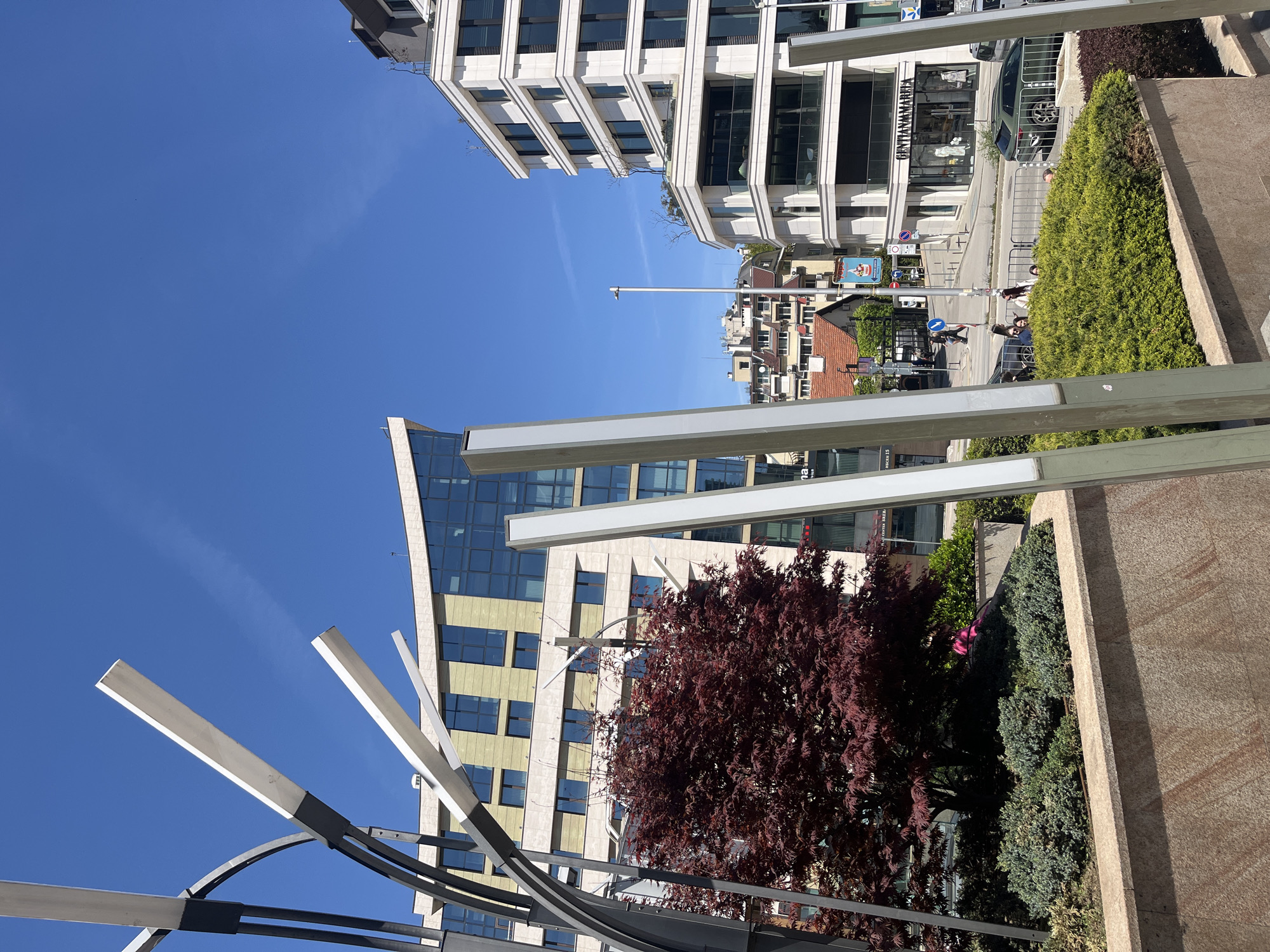 The image depicts an outdoor scene in Sofia, Bulgaria, under a clear blue sky. The foreground includes modern architectural elements, specifically a metal structure with white rectangular bars extending upwards. Adjacent to this is a landscaping feature with a red-leaved tree and various green shrubs.
In the background, buildings of varying architectural styles are visible. One is a contemporary structure with a glass facade and tan-colored panels, while another appears to be a white building with repetitive window patterns.
A street scene is partially visible, with pedestrians crossing at a crosswalk, and a car parked nearby. A storefront labeled "GENTIANAMARKET" can be seen on one of the buildings. Street signs are also present, indicating street directions and restrictions.