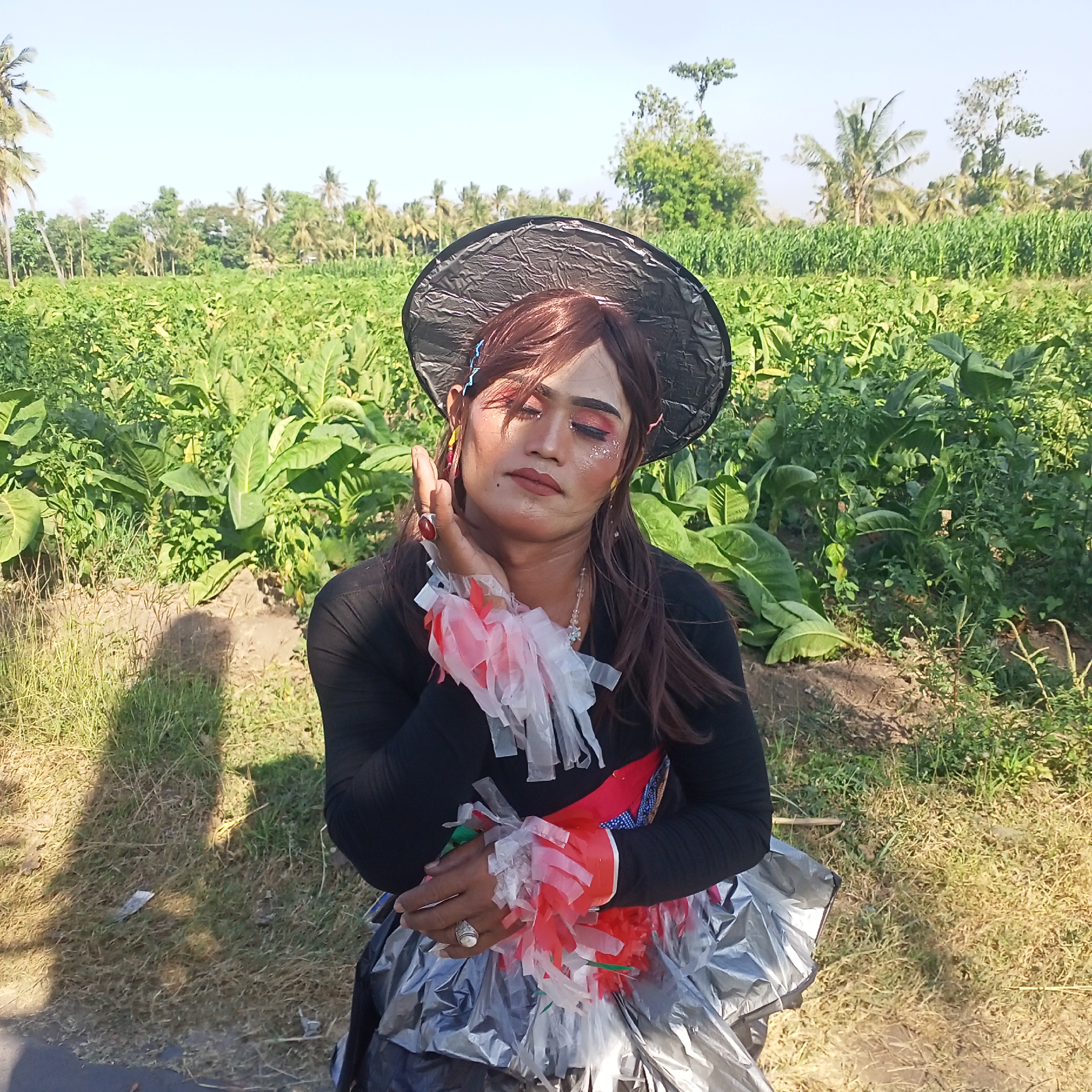 The image depicts a person with makeup, a wig, and a hat, posing in what appears to be a tobacco field in Kedung Dowo, Indonesia. The person is wearing a black long-sleeved shirt adorned with red and white fabric streamers on the sleeves and a large, shiny skirt made of silver and black material. The individual's face is partially obscured by their hand, which is decorated with rings and streamers. The background consists of rows of lush green tobacco plants under a clear blue sky, with scattered palm trees and other vegetation visible in the distance. The ground is a mix of dry soil and grass. The lighting suggests it is daytime, possibly late morning or early afternoon, with a shadow cast on the ground to the left of the subject.