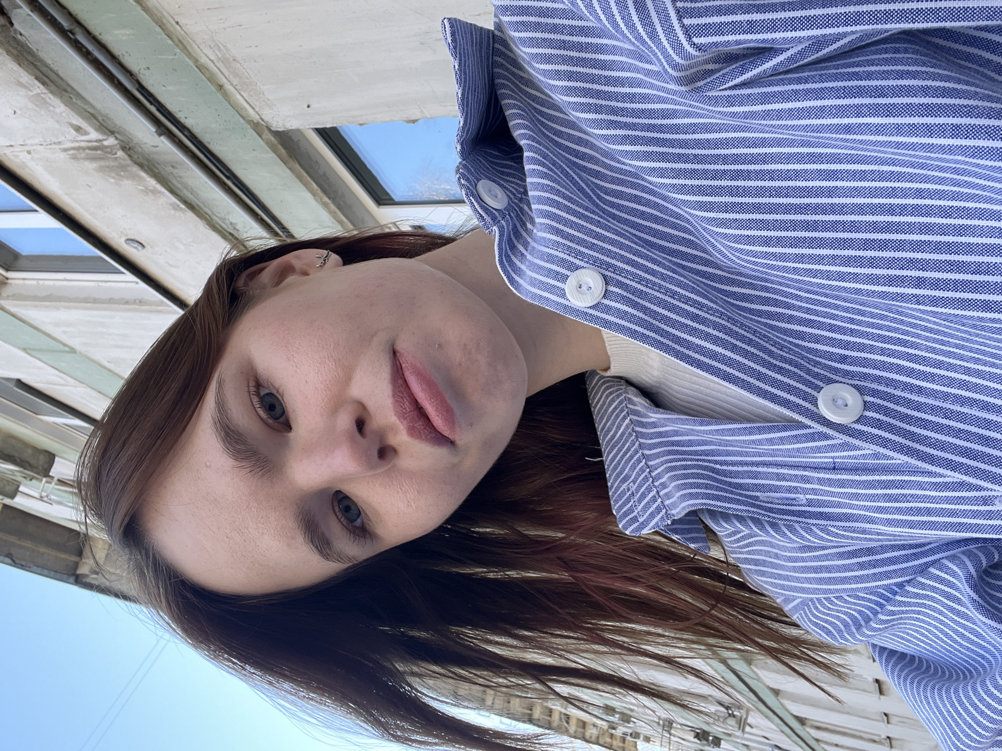 A woman is depicted from a low angle, with her head tilted to the side. She has fair skin, light eyes, and brown hair with reddish highlights near the ends. She is wearing a blue and white striped button-up shirt over a white underlayer. A small silver earring is visible in her left ear.
In the background, the blurry facade of a building is visible. Given the location constraint of Chaikovsky, Russia, it is likely that this building is within the city limits. Details of the building's architecture are not distinctly visible, but some window frames and structural elements suggest a multi-story building. The sky is visible behind and to the side of the building, indicating an outdoor setting.