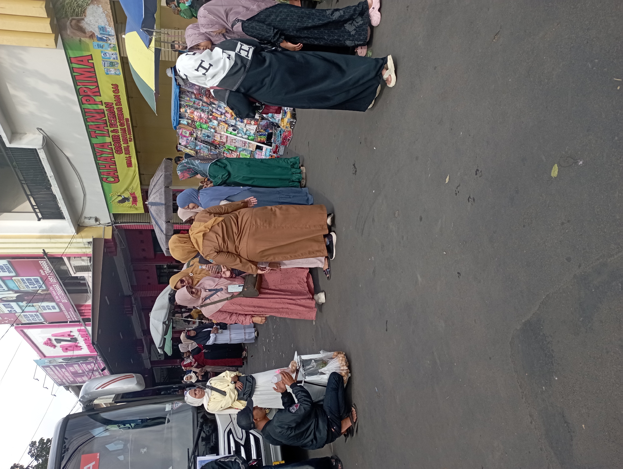This is a medium shot, eye-level photograph taken outdoors on a street, likely in the city of Tasikmalaya, Indonesia, given the context of the text on the signs. The scene depicts a busy marketplace or roadside stall area with several people engaged in various activities.
In the foreground on the left, a vendor wearing a black jacket and a black baseball cap is crouching down, interacting with a customer. He is attending to a small white cart filled with packaged goods, possibly snacks, and has a plastic bag with more items in it. The customer, a person in a light yellow hijab, stands to his right, appearing to be making a purchase.
Behind the vendor, a large grey bus is parked, partially visible on the left side of the frame. The bus has tinted windows and a reflective silver trim.
The middle ground of the image is populated by a group of people, mostly women wearing hijabs and modest clothing in various colors like pink, beige, green, and blue. They are standing and conversing. To the right of this group, a stall is set up with an array of colorful products displayed on shelves. Umbrellas of red, yellow, and green are providing shade over the stalls.
In the background, buildings with signage are visible. One prominent sign in green and yellow text reads "CAHAYA TANI PRIMA" with smaller text below indicating "GROSIR & ECERAN" and "BERAS, GULA, MINYAK GORENG DAN GAS." Other signs are also present, some with Indonesian text and graphics. The architectural style suggests a commercial area. The ground is paved with asphalt, showing some signs of wear and tear. The overall atmosphere appears to be that of a lively street market on a bright, possibly sunny day.