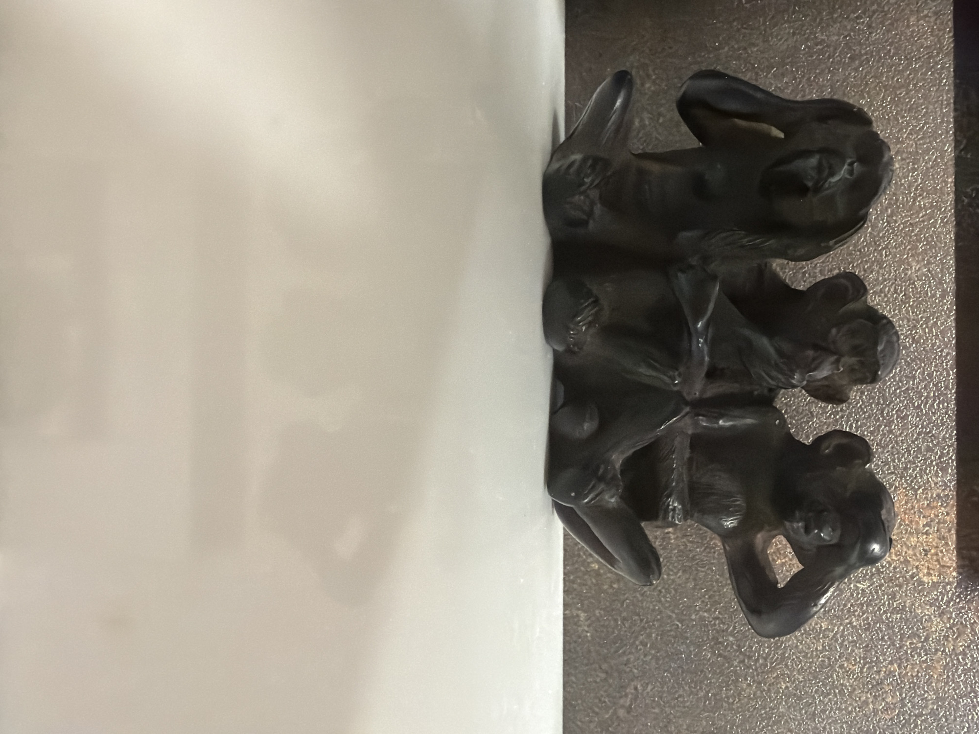 The image shows a decorative element consisting of three stylized animal figures stacked vertically, likely made of dark metal. The figures appear to be stylized depictions of cats or similar creatures. The element is attached to the edge of a white object, possibly a piece of furniture or architectural feature. The background suggests a surface with a textured or granular appearance. The photograph was taken in Arinj, Armenia.