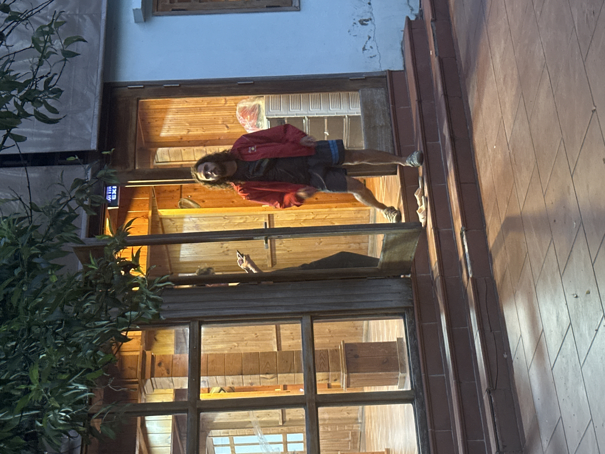A person is standing in the doorway of a building, Du Già Commune, Vietnam. The person has long hair and is wearing a red jacket and shorts. The building has a wooden frame and glass windows. There is a bush to the left of the doorway and tiled stairs to the right. Inside the building, another person is visible in the background holding a phone.