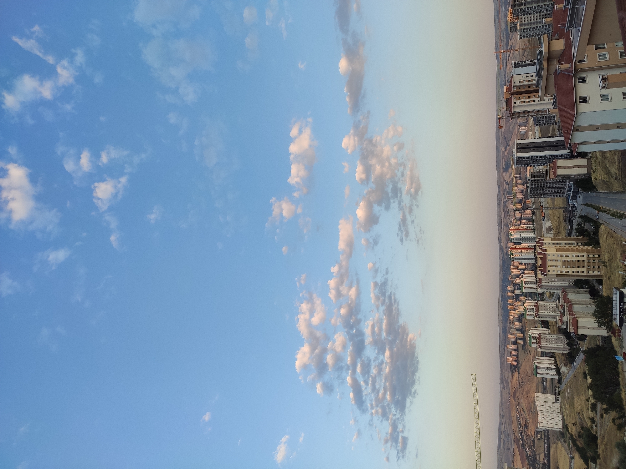 An aerial, wide shot captures a vast cityscape with a prominent presence of residential buildings and construction cranes under a clear, partly cloudy sky. The scene is set in Etimesgut, Turkey. The sky transitions from a vibrant blue overhead to a pale yellow and pink hue near the horizon, indicating either early morning or late evening. Scattered white and grey clouds dot the sky, some tinged with the warm light of the setting or rising sun.
Dominating the middle and lower portions of the image is a dense cluster of apartment buildings. These structures vary in height and architectural style, with many featuring red-tiled roofs and balconies. Some buildings display a mix of concrete, white, and brown facades, while others are darker, with grey and black exteriors. A few buildings appear to be under construction, evidenced by visible scaffolding and the presence of large yellow construction cranes scattered throughout the urban landscape. One particularly prominent crane arm extends horizontally across the left side of the frame, silhouetted against the sky.
The urban sprawl extends into the background, where more buildings are visible on rolling hills. The terrain surrounding the city is arid and brown, with sparse vegetation. A road winds through the development in the foreground, with a few small vehicles visible. The overall impression is one of a rapidly developing residential area on the outskirts of a city. No people are discernible in the image. The environment is dry and dusty, characteristic of a semi-arid climate.