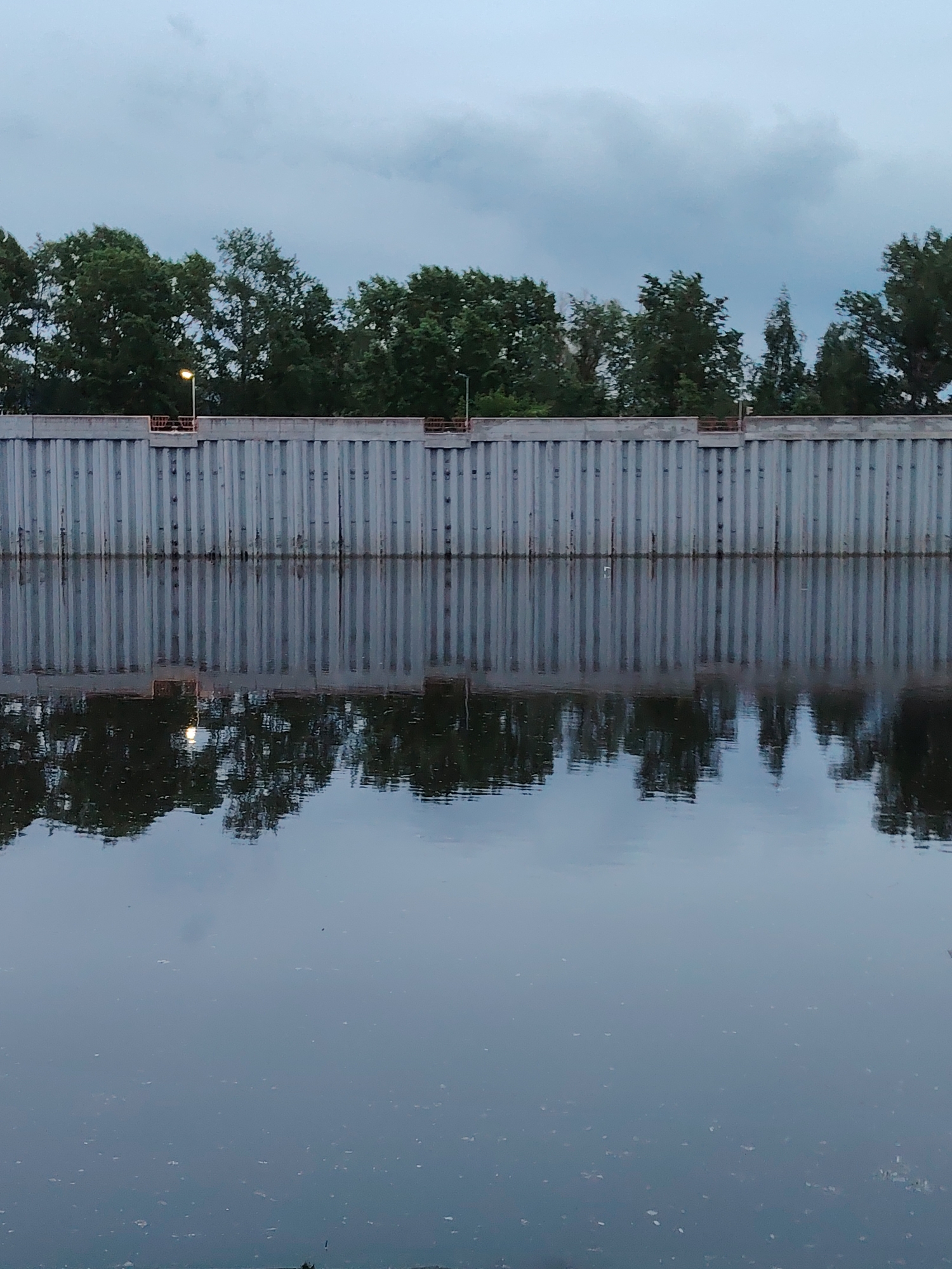 The image depicts a body of water, likely a river or canal, reflecting a gray, corrugated wall. The wall is topped with trees and a lamppost. The sky is overcast and gray. The location is Chaikovsky, Russia. There are no people visible in the image.