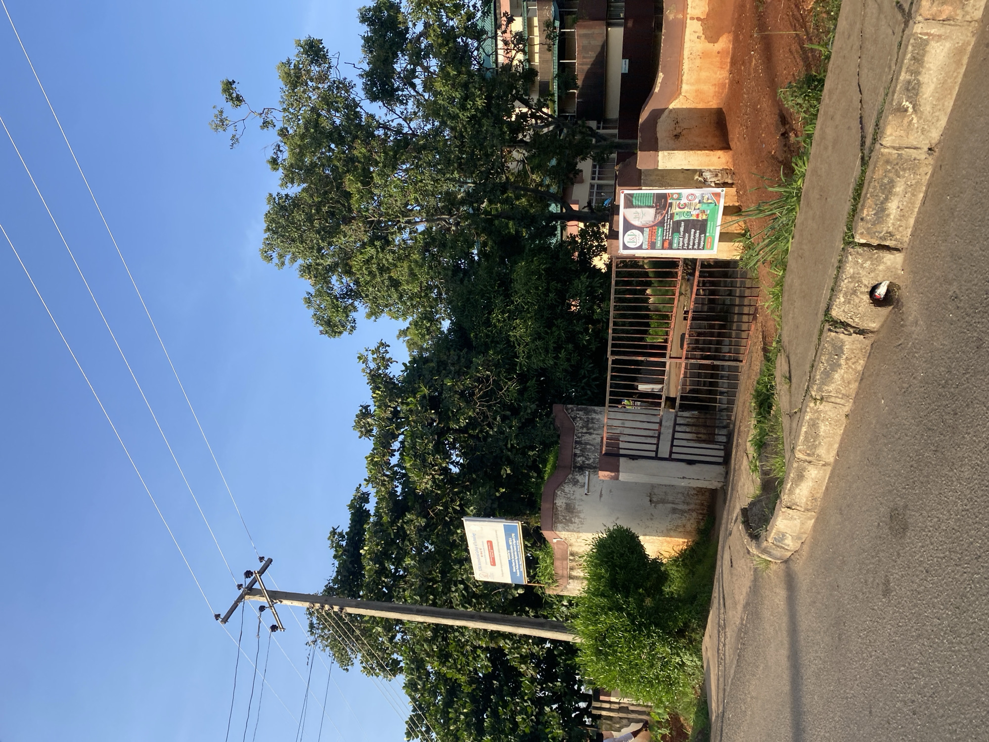 The image depicts a street view in Jos, Nigeria, on a clear, sunny day. In the foreground, a paved road curves to the left, with a concrete curb and a drainage ditch. To the right of the road, a section of a property wall is visible, painted in earthy tones of orange and brown. A black metal gate stands partially open, leading into the property. A signpost on the wall displays an advertisement with "J&J" prominently featured. Behind the wall and gate, lush green trees and foliage dominate the scene. A tall wooden utility pole with multiple electrical wires stretches across the sky. In the middle ground, a white sign with blue lettering is attached to a concrete structure, reading "ESCA International College of Technology." Further back, a multi-story building with a green roof is visible, partially obscured by trees. The overall environment suggests a tropical or subtropical climate.