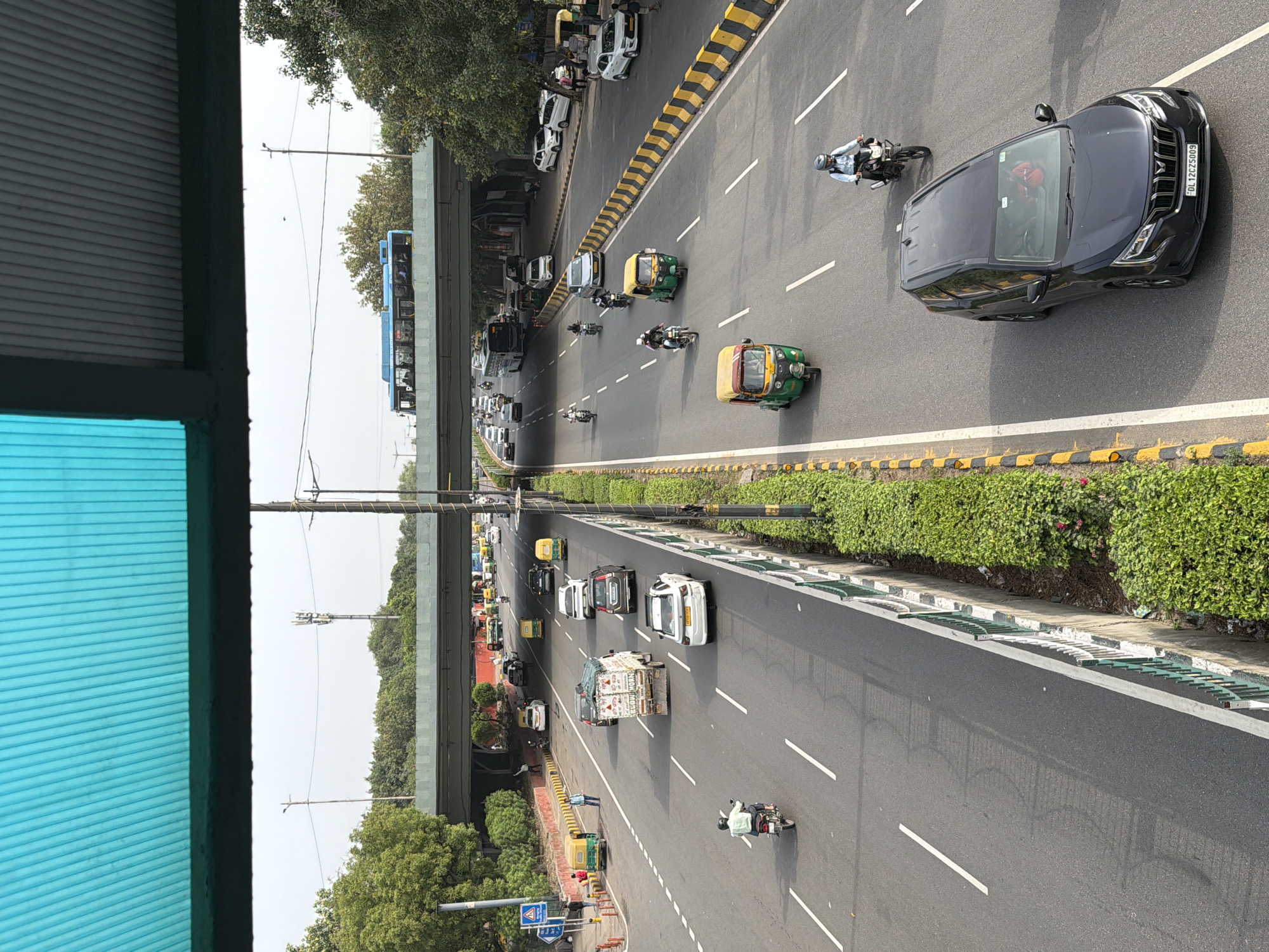 The image shows a busy multi-lane road in Delhi, India, with vehicles including cars, motorcycles, auto-rickshaws, and a truck. The vehicles are traveling in both directions, separated by lane markings and a central divider with a green metal railing and dense green foliage. An overpass is visible in the background, with a blue bus crossing it, and trees line the sides of the road. In the foreground, there is a dark-colored SUV with the license plate "DL 12CZ5009". The scene appears to be a typical urban street with moderate traffic and pedestrian activity on the sidewalks.